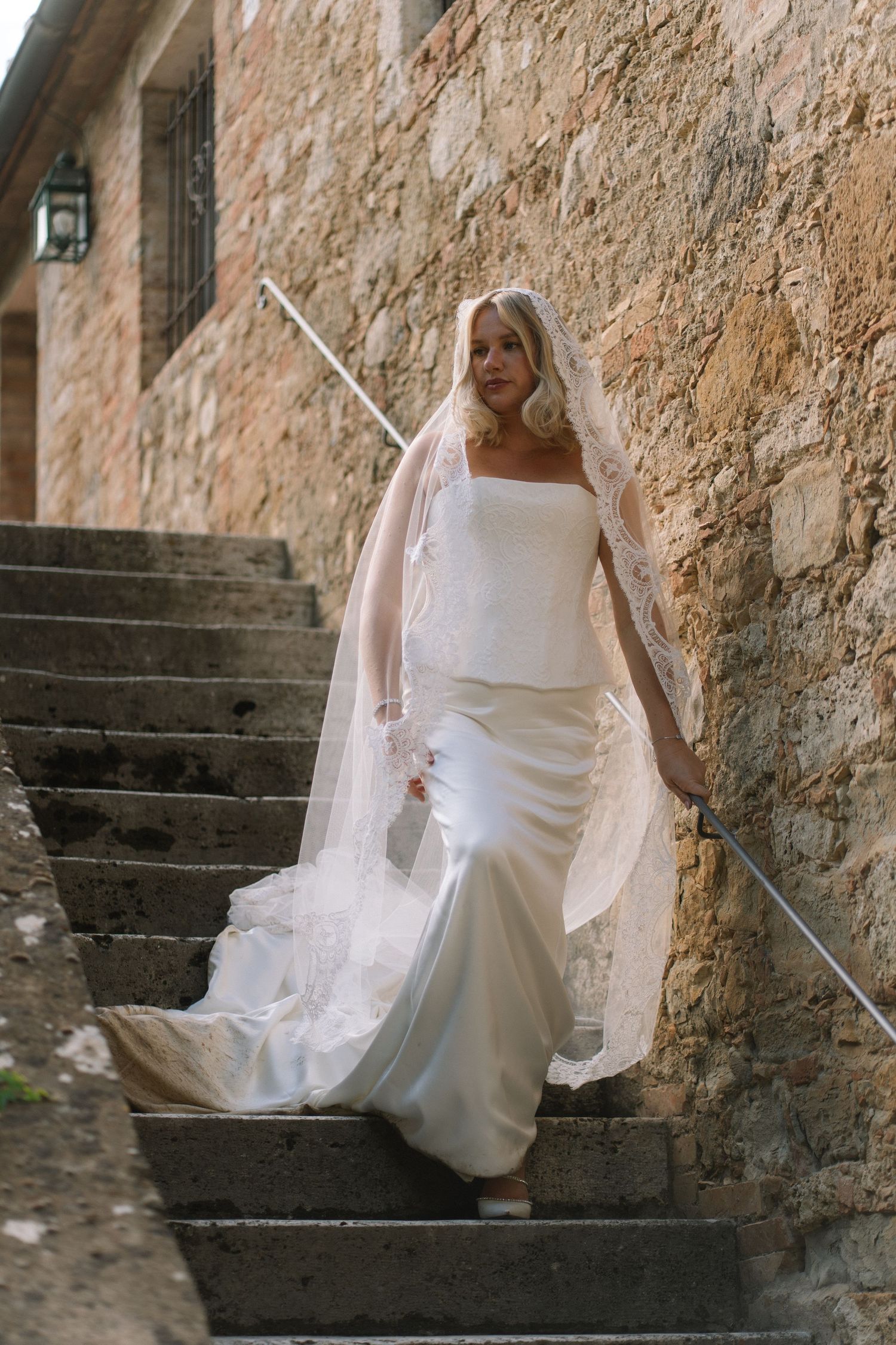 Stunning blonde bride in white satin mermaid gown and lace veil walking down stone steps at Tuscan venue.