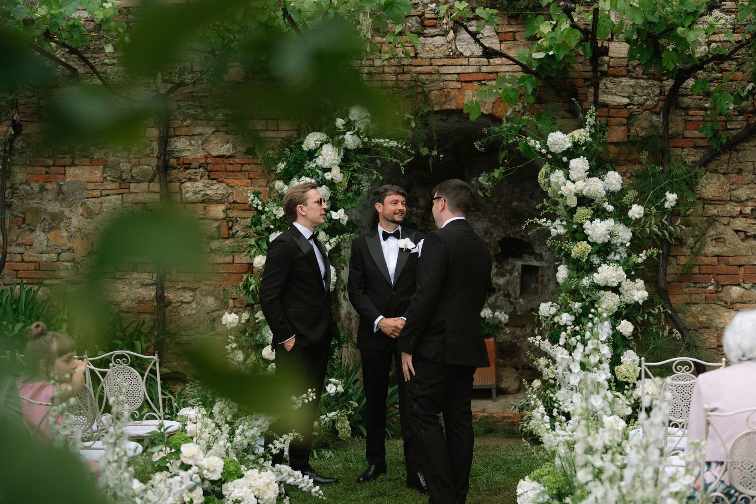 Two grooms in black tuxedos standing at a floral arch during an outdoor garden wedding ceremony.