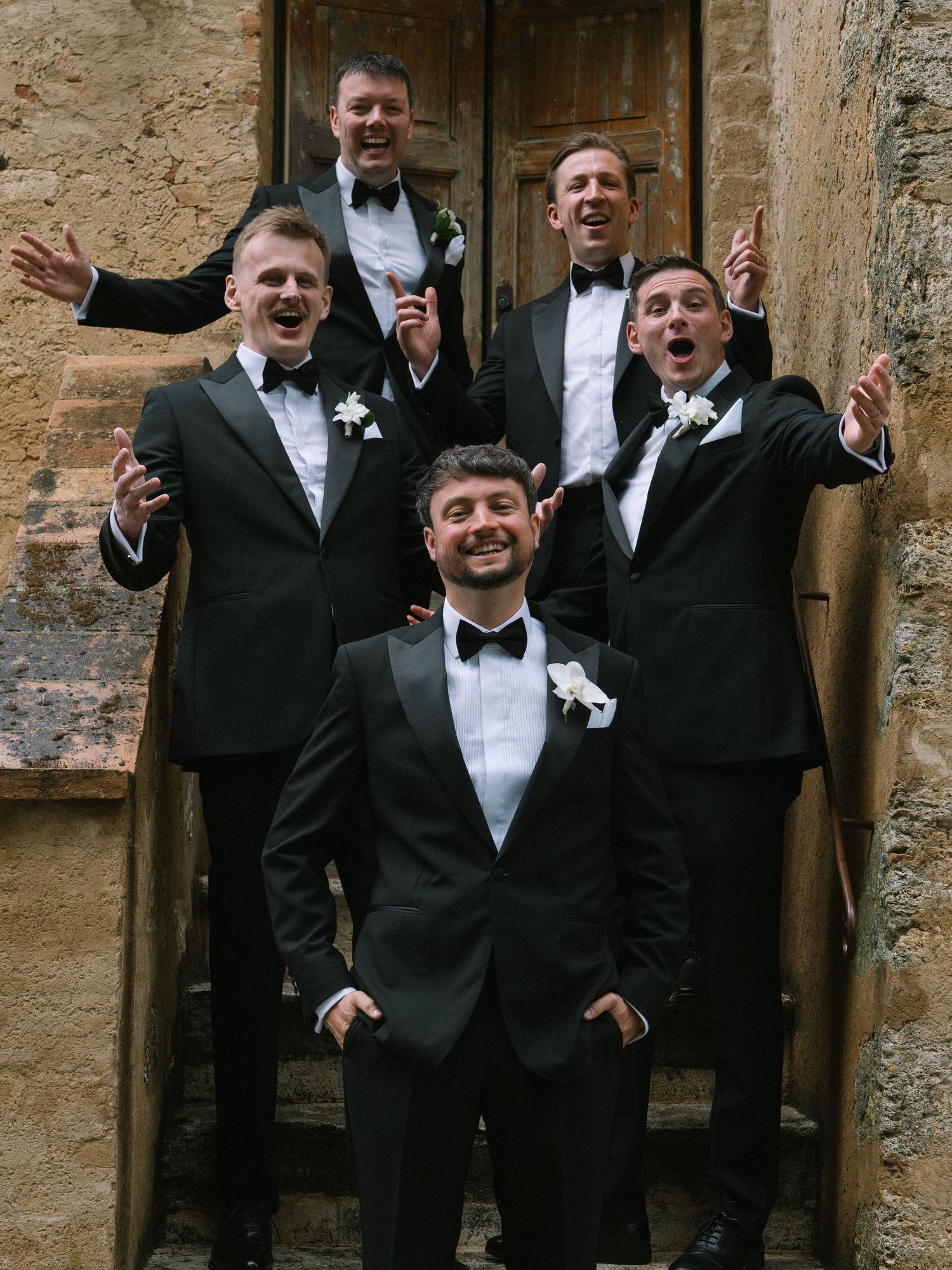 Five groomsmen in black tuxedos celebrating joyfully on stone steps of rustic Italian venue.