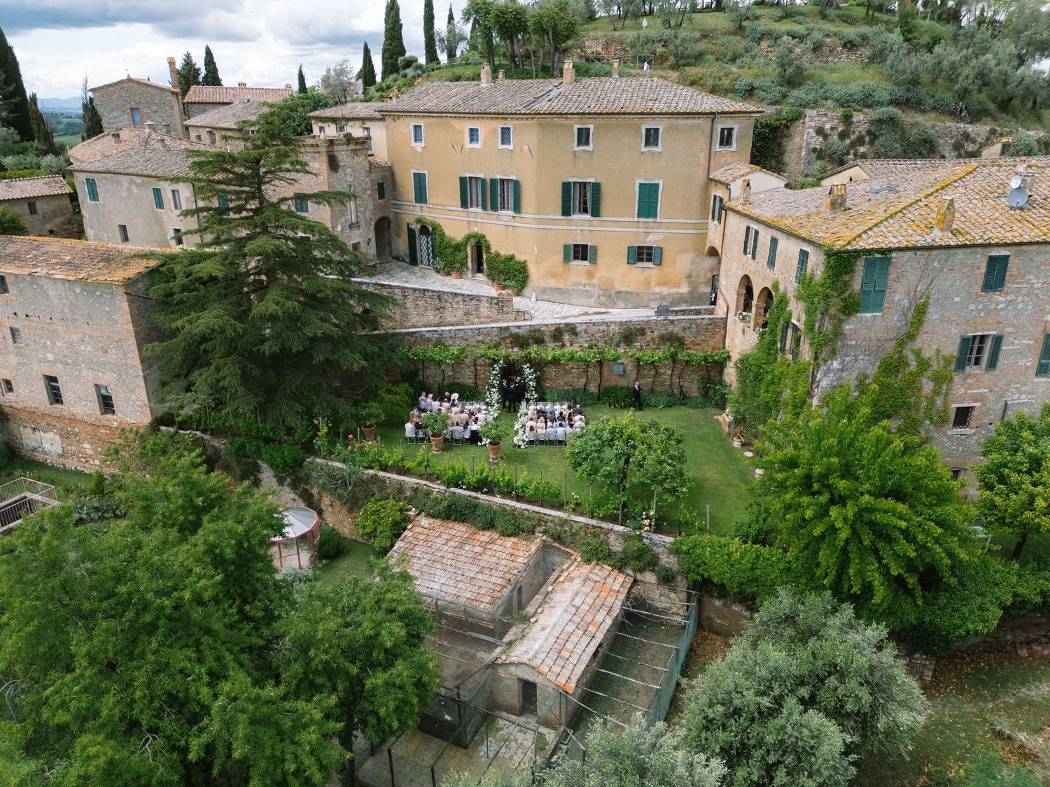 Aerial view of a Tuscan villa wedding ceremony with guests seated in a lush garden courtyard surrounded by stone buildings.
