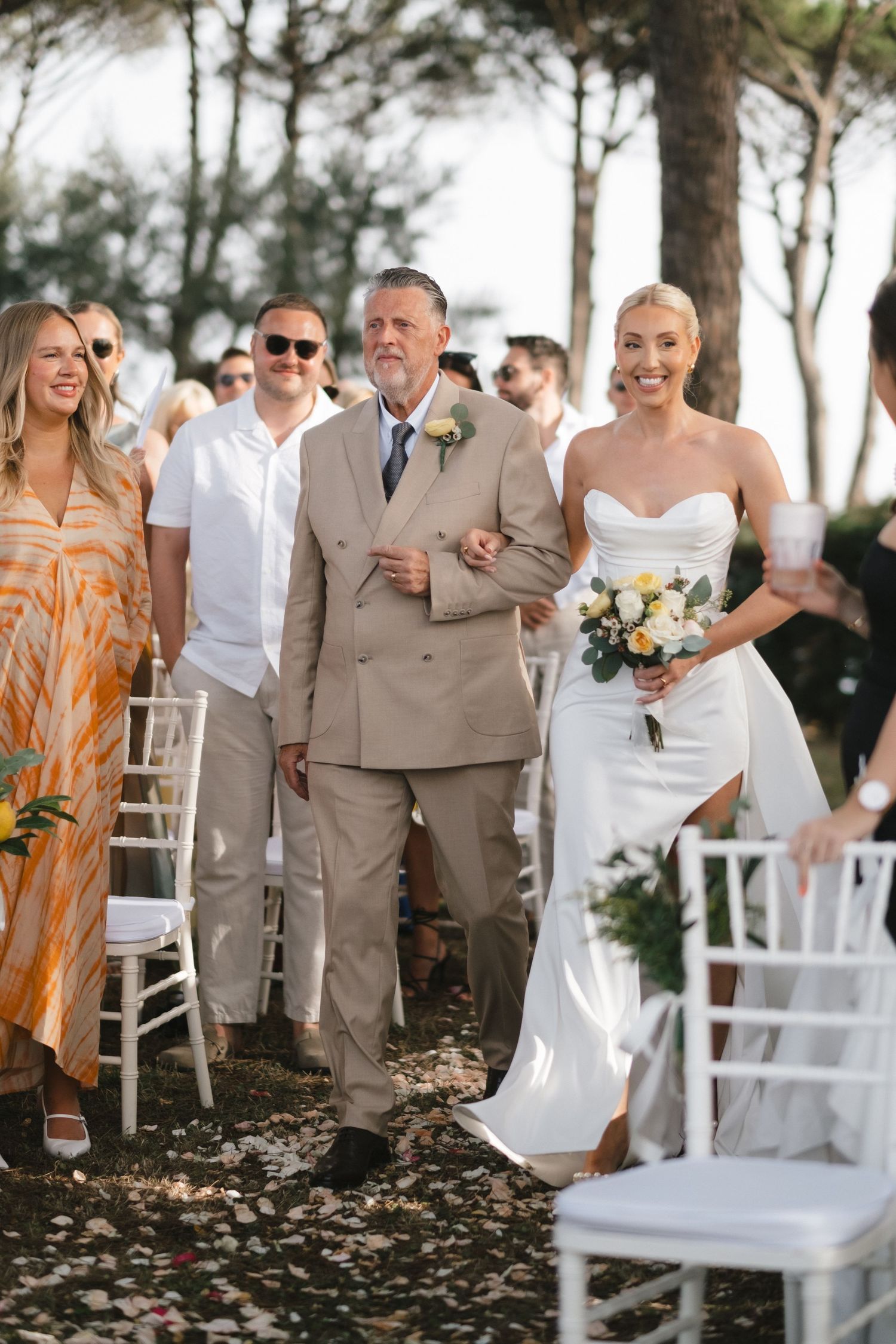 Bride in white gown walks down outdoor aisle with father, holding yellow and white bouquet at garden wedding ceremony.