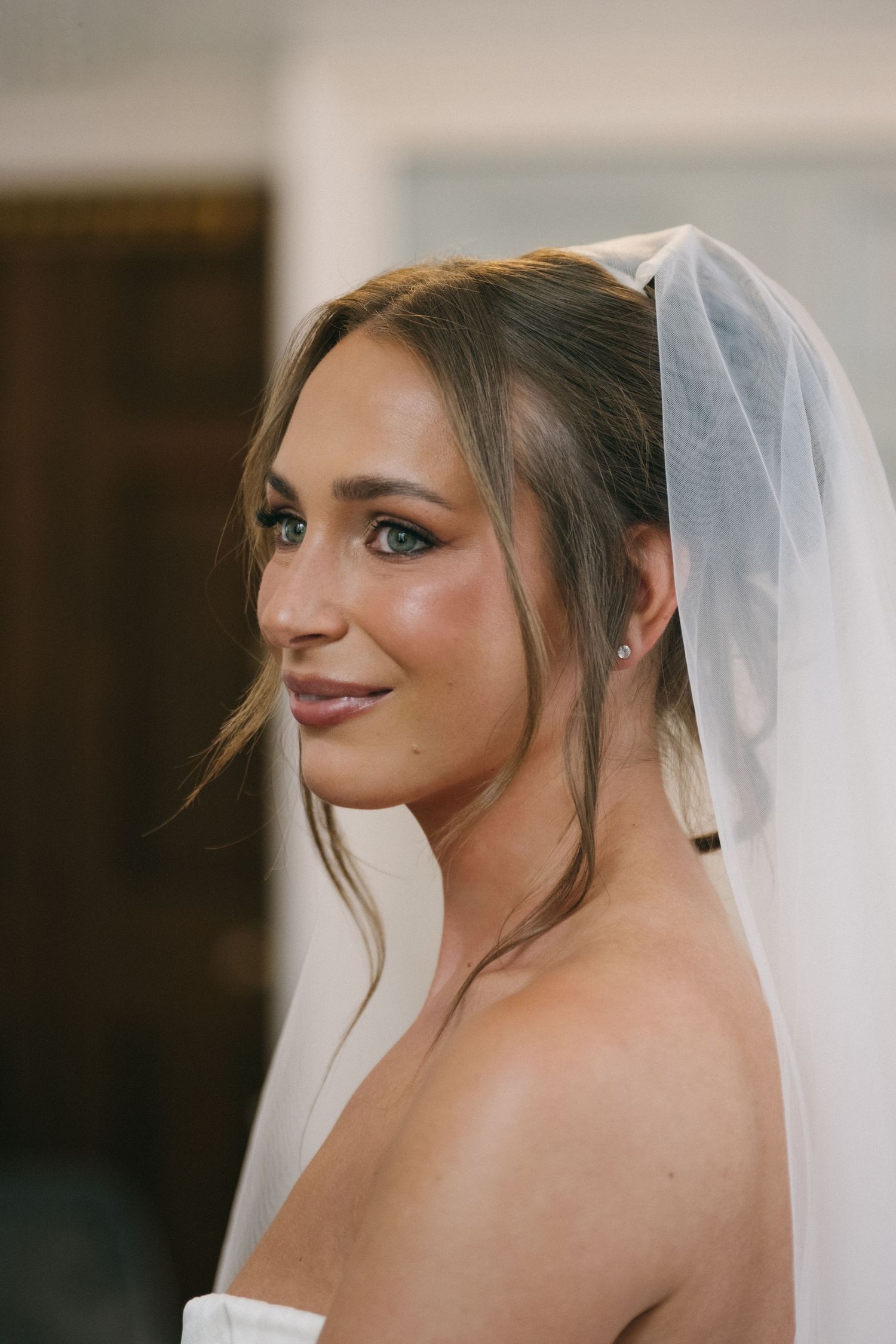A beautiful bride in a white strapless gown and sheer veil smiles softly over her shoulder in an elegant portrait.