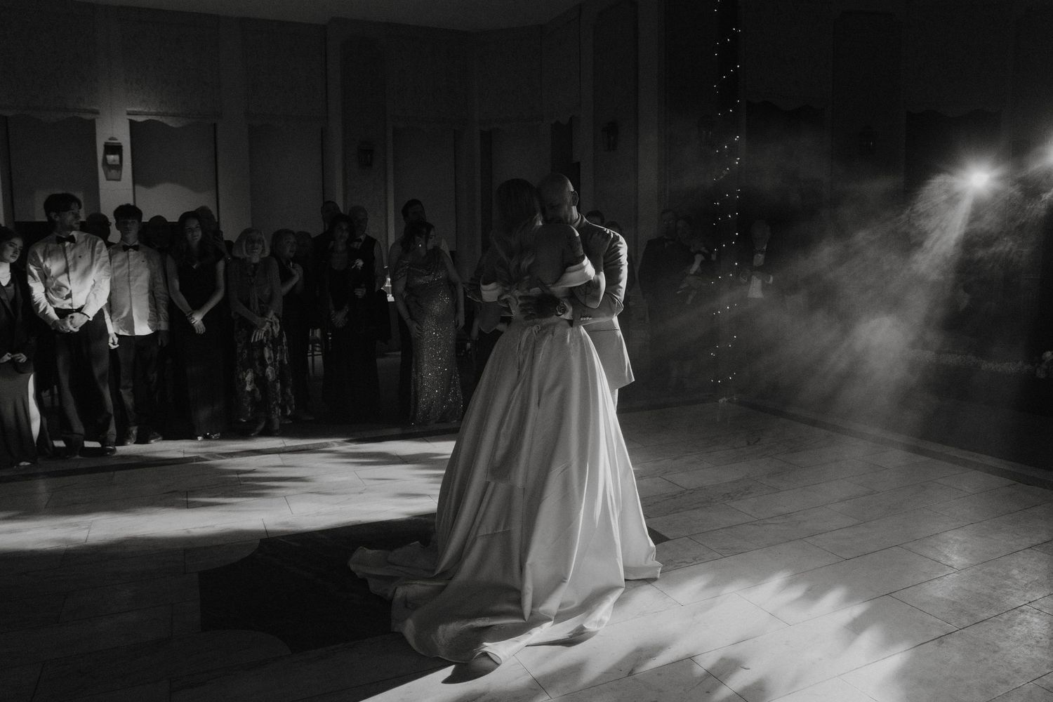 Black and white photo of a couple sharing their first dance at a wedding reception with guests watching in the background.