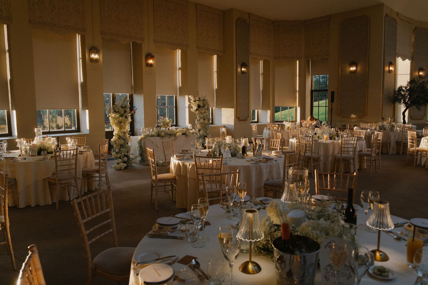 Elegant wedding reception hall with round tables, gold chiavari chairs, white linens, and candlelit centerpieces.