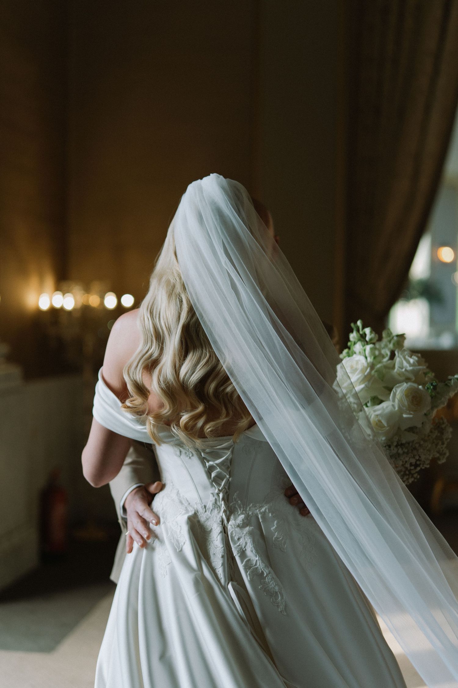 Bride in white off-shoulder gown with long veil holding bouquet, back view in elegant candlelit room.