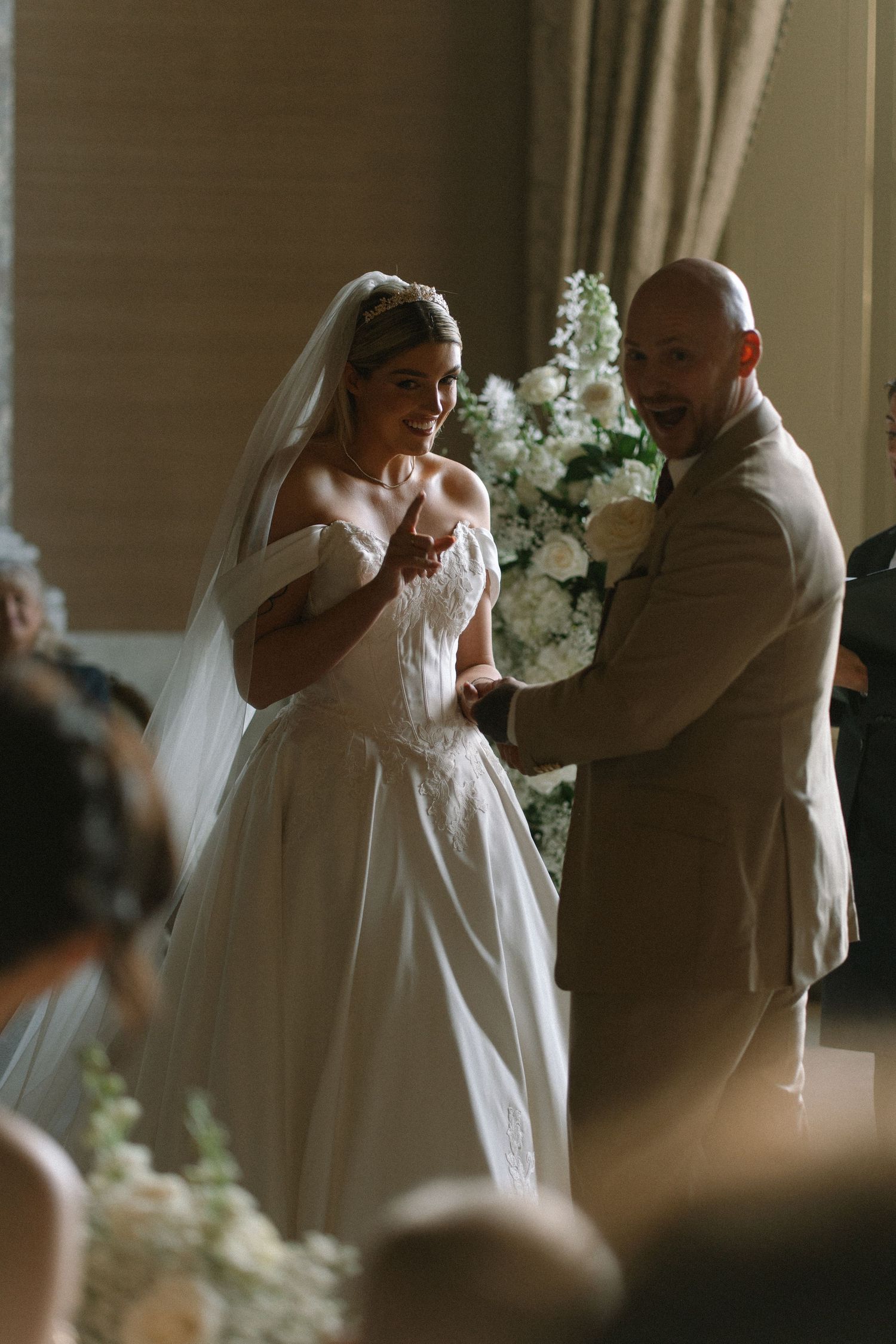 Bride in white gown and veil exchanges rings with groom in tan suit at elegant wedding ceremony with white floral backdrop.