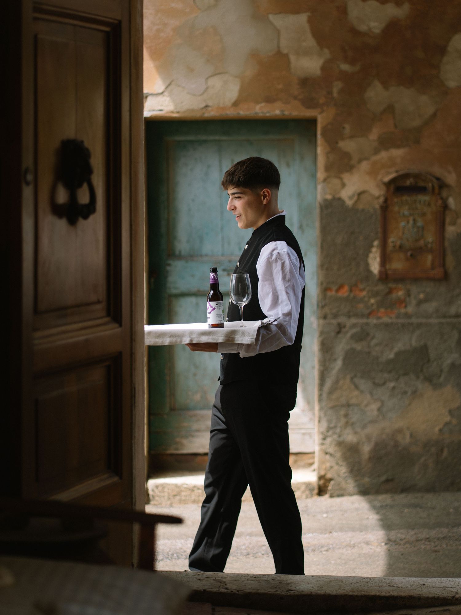 Young waiter in black vest carrying wine tray through rustic doorway at wedding venue.