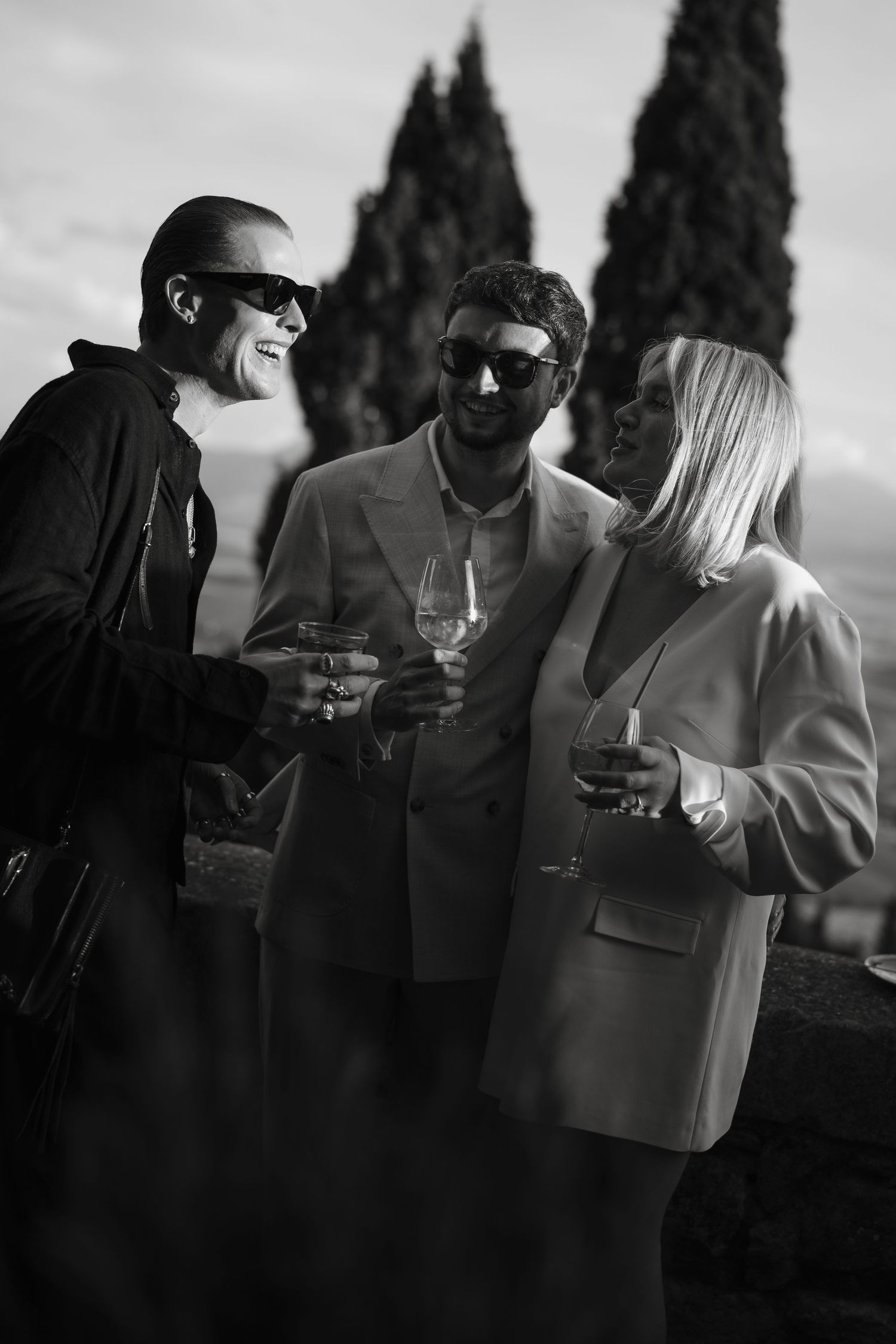 Black and white photo of three stylish wedding guests in sunglasses enjoying champagne at an outdoor Tuscan celebration.