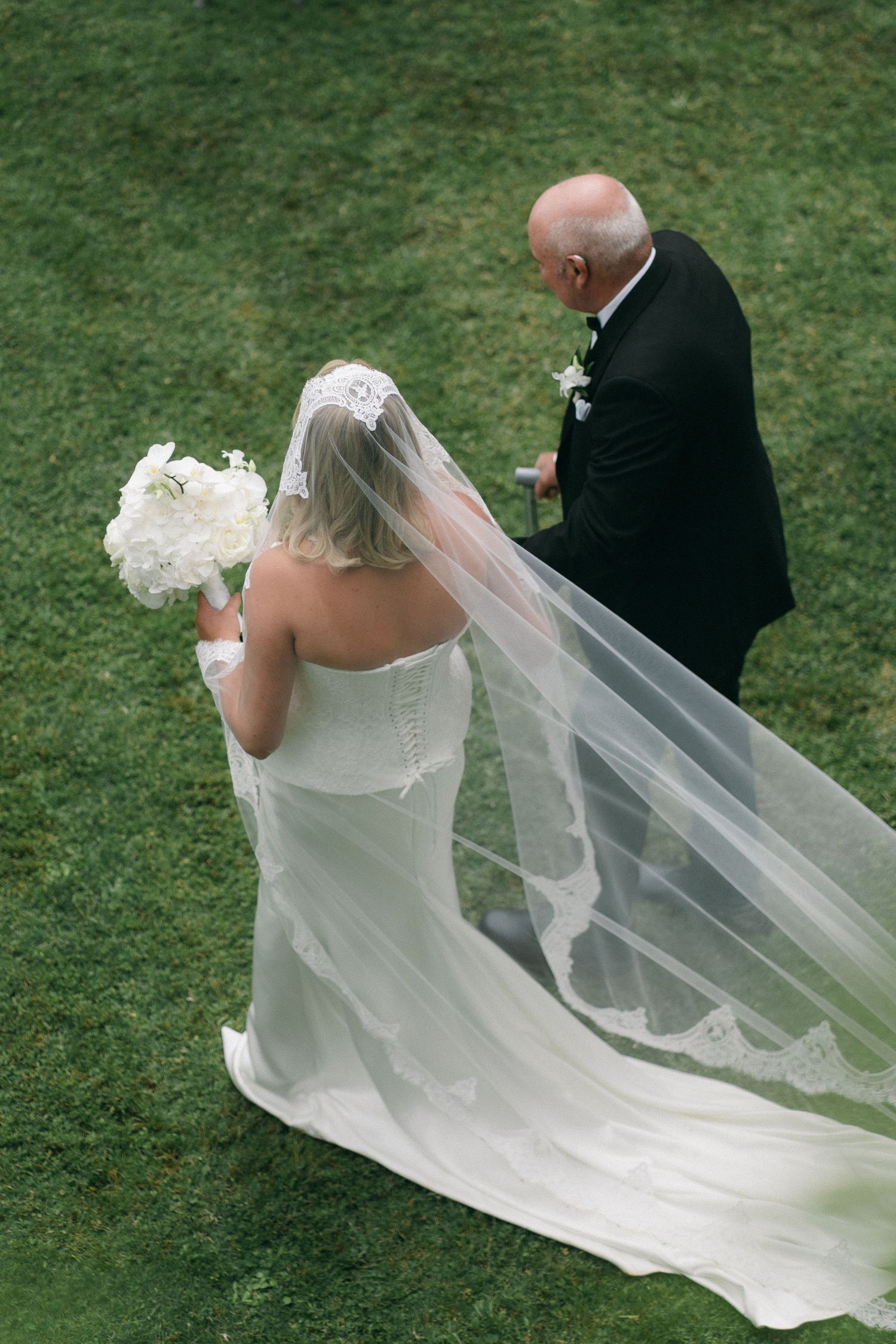 Aerial view of bride in white gown with cathedral veil being escorted by bald man in black tuxedo on green lawn.