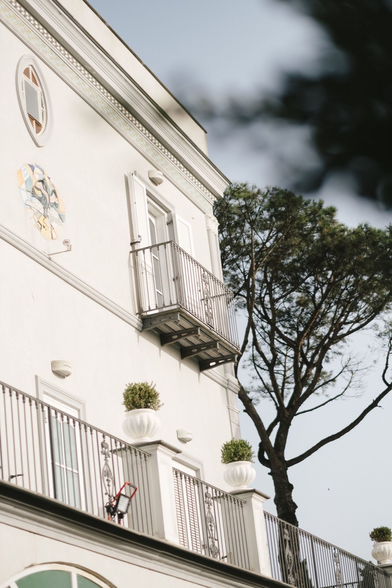 Elegant white Italian villa facade with ornate balconies, decorative crest, shuttered windows, and Mediterranean pine tree.
