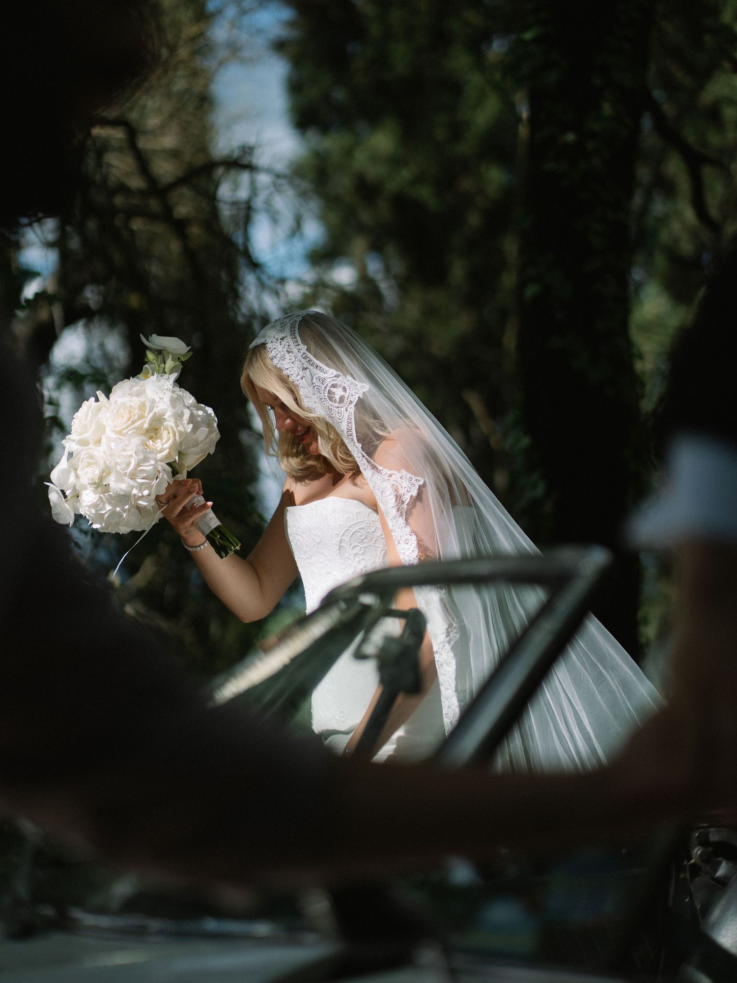 Elegant bride with lace veil holds white peony bouquet beside a classic vintage wedding car outdoors.