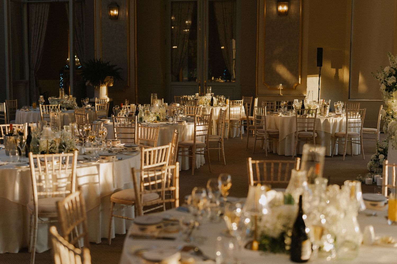 Elegant wedding reception hall with gold chiavari chairs, round tables, white linens, and soft ambient lighting.