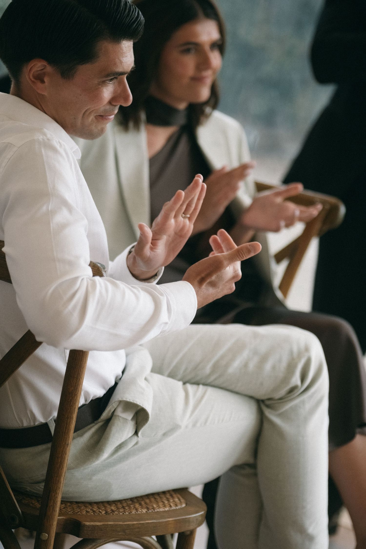 Man in white shirt gestures with hands while seated at a business meeting with colleagues in background.