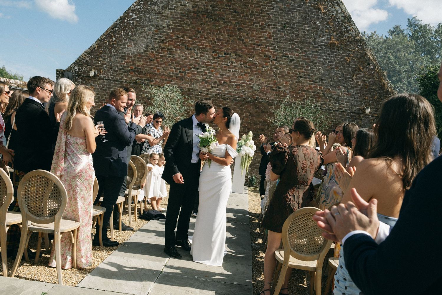 Bride and groom share a kiss at outdoor wedding ceremony aisle, guests applauding beside rustic brick barn.