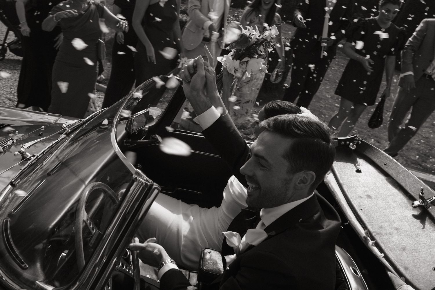 Black and white wedding photo of groom waving from vintage car as confetti falls around the couple.