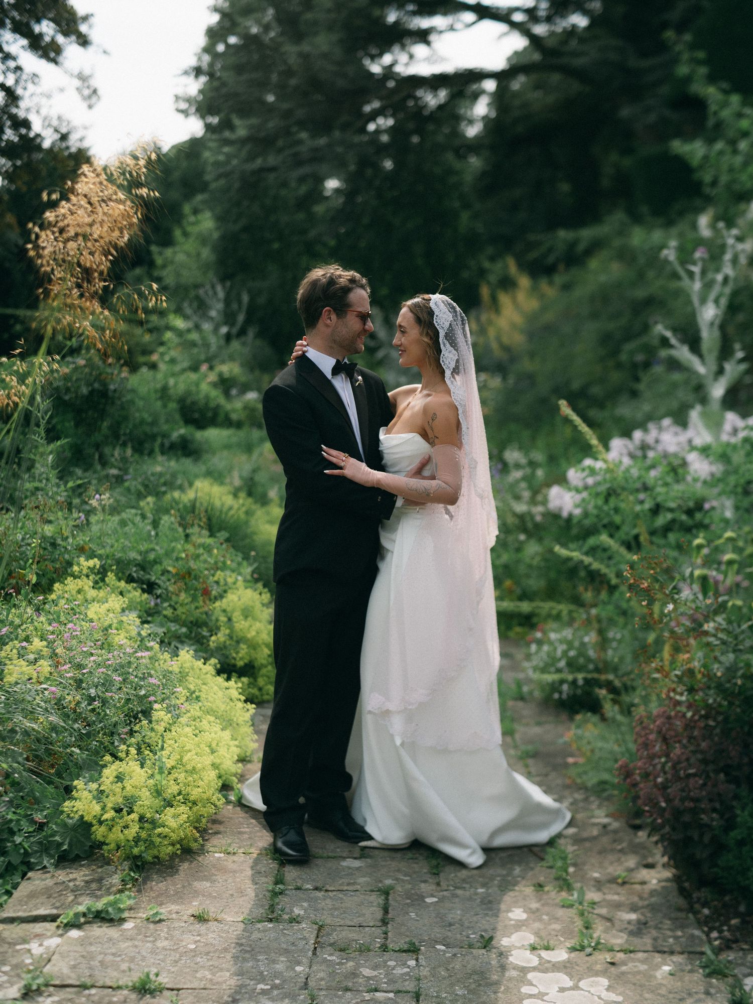 A bride and groom share a romantic moment in a lush garden, she wearing a white gown and veil, he in a black tuxedo.