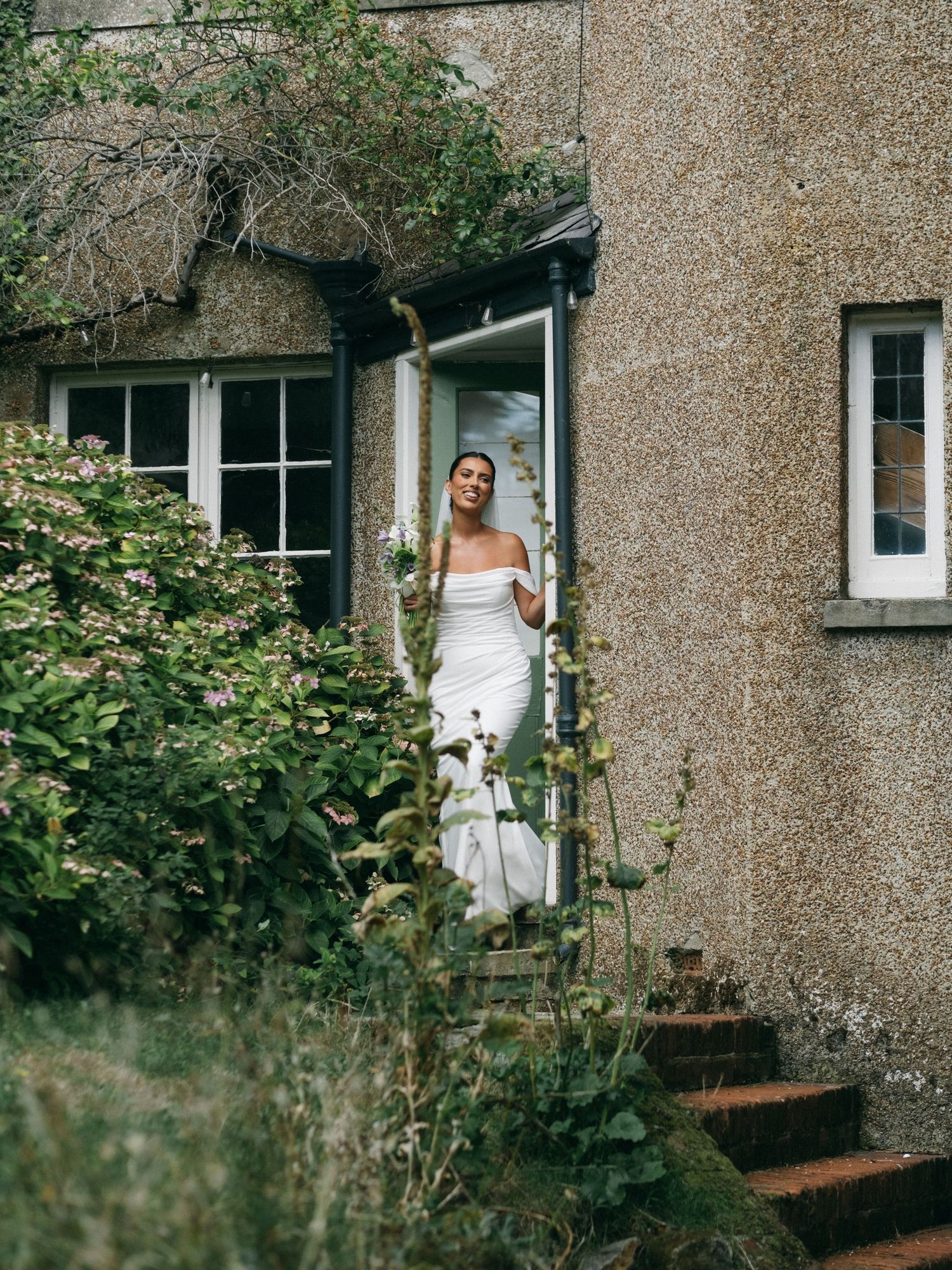 Bride in off-shoulder white gown stands in doorway of rustic stone cottage surrounded by lush greenery.