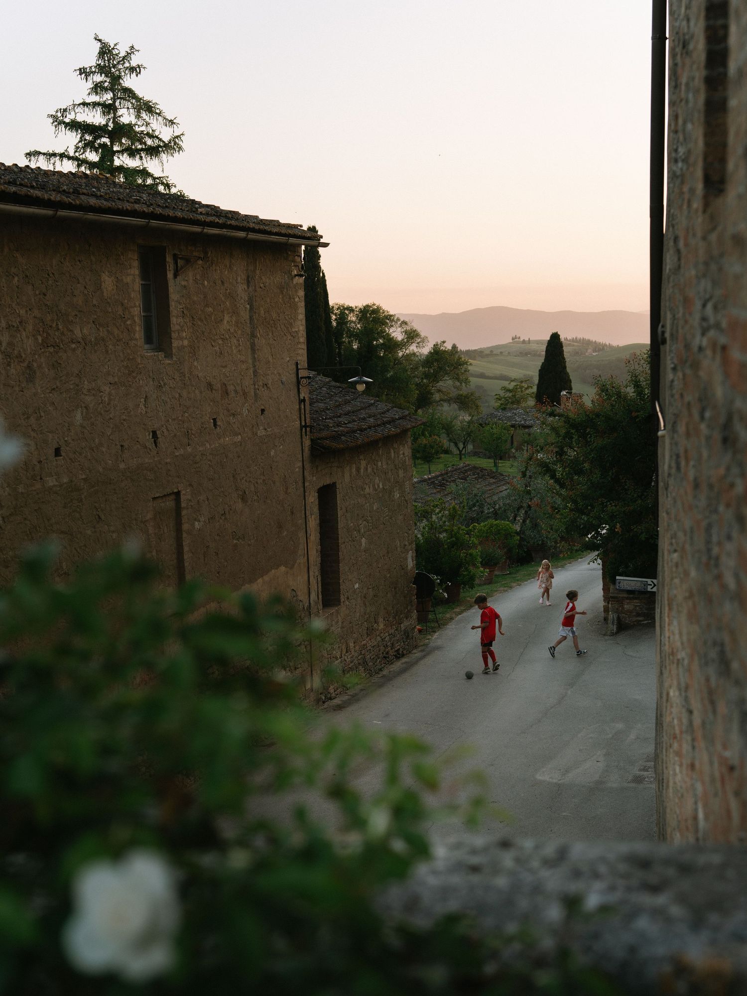 Children playing on a narrow cobblestone street between rustic stone buildings in a Tuscan village at sunset.