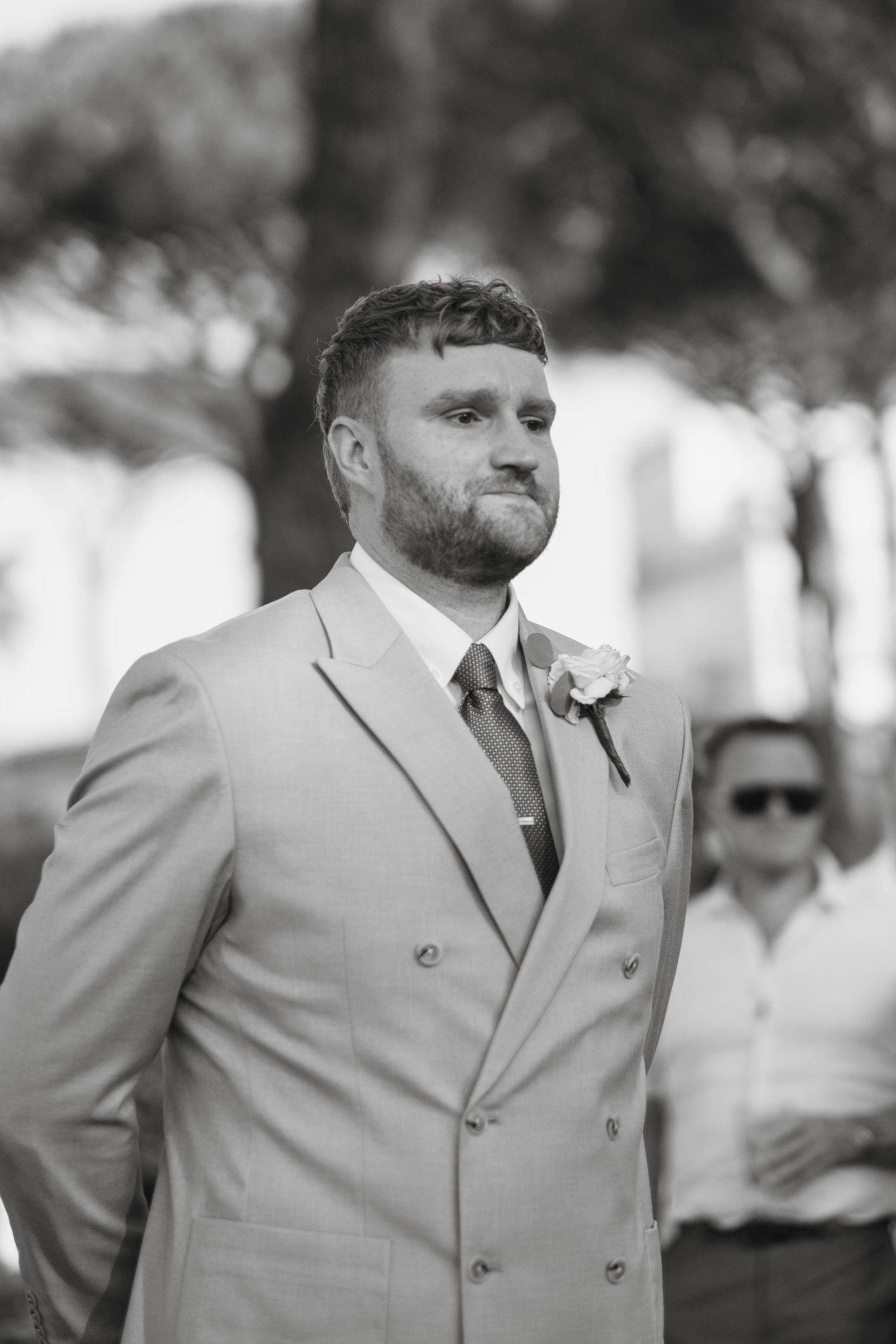 Black and white portrait of bearded groom in tan double-breasted suit with boutonniere at outdoor wedding.