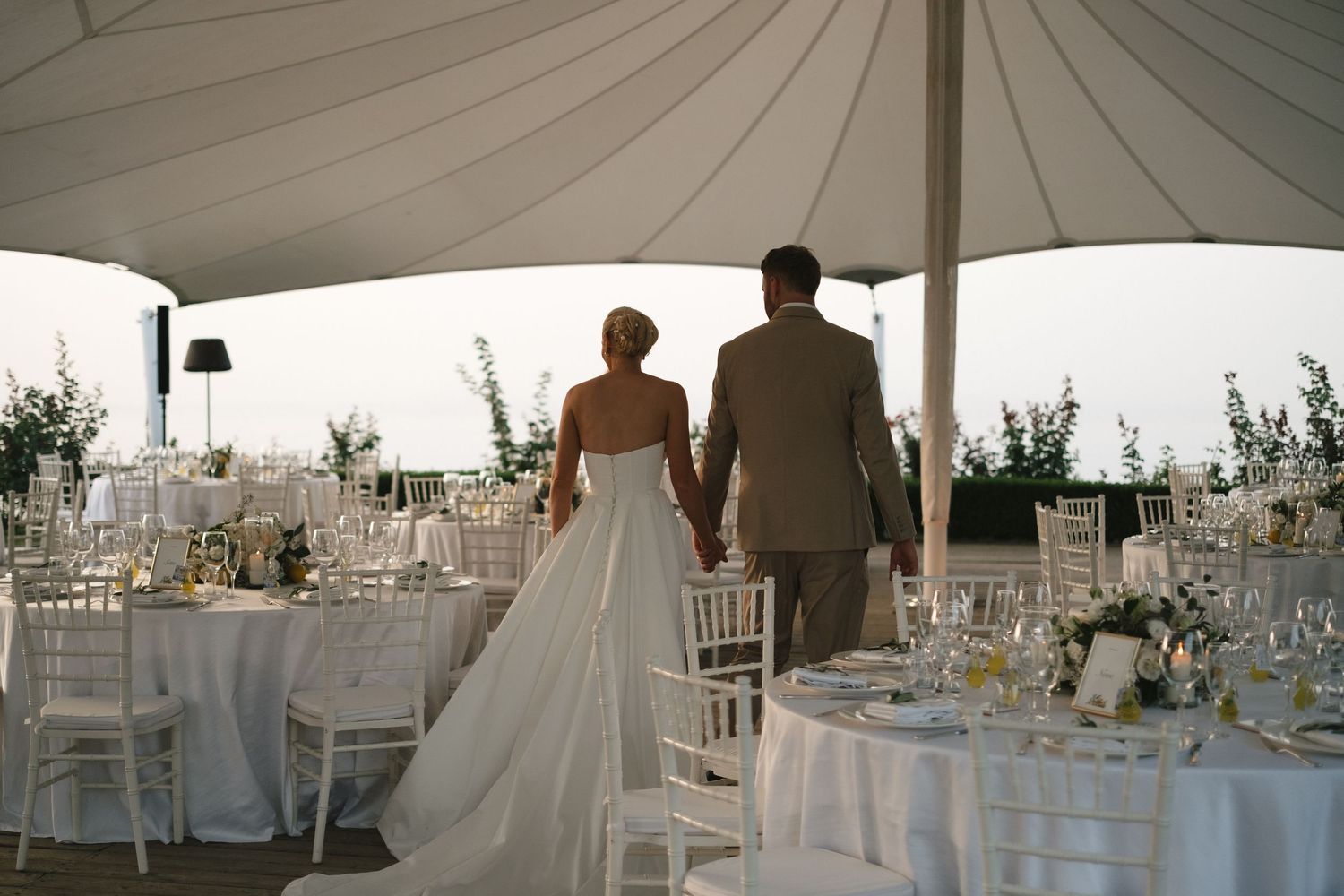 Bride and groom holding hands viewing their elegant outdoor wedding reception venue under a white tent.
