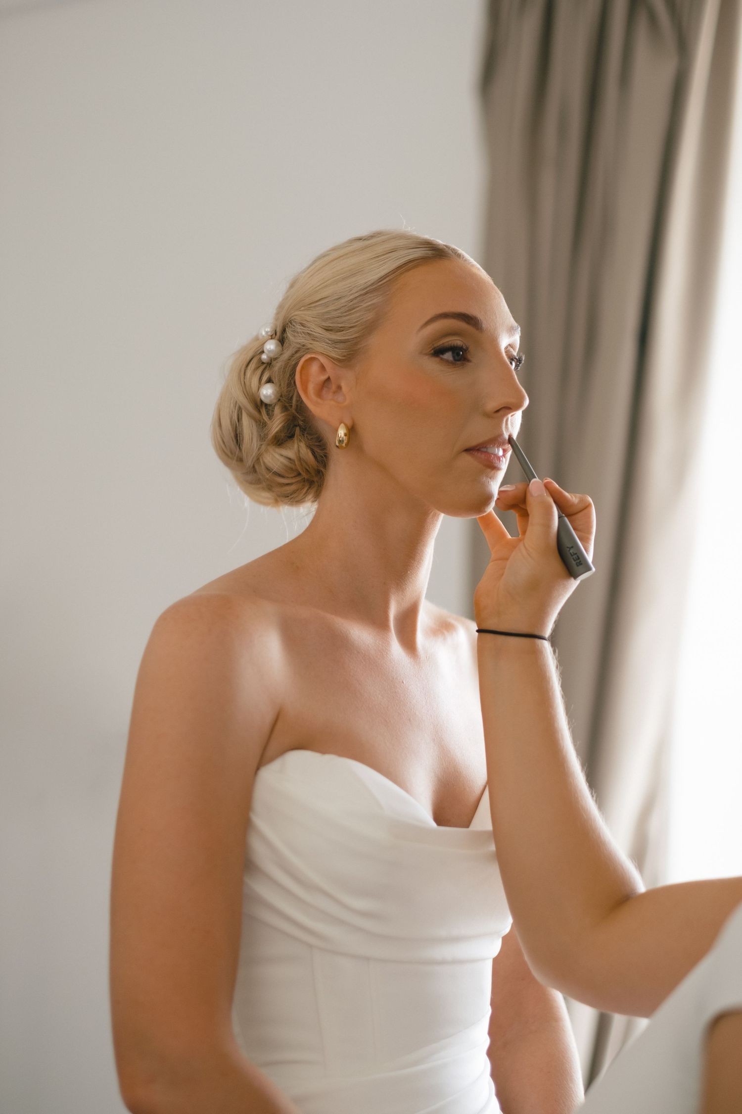 Bride in strapless white gown gets lipstick applied by makeup artist, hair styled in elegant updo with pearl pins.