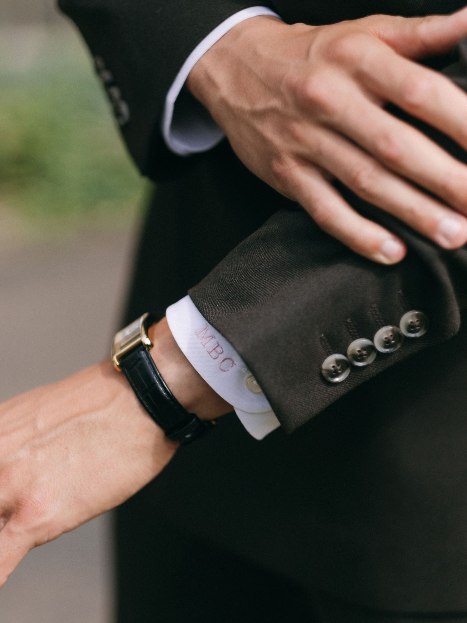 Close-up of groom's wrist wearing gold watch with black leather strap against dark suit sleeve.