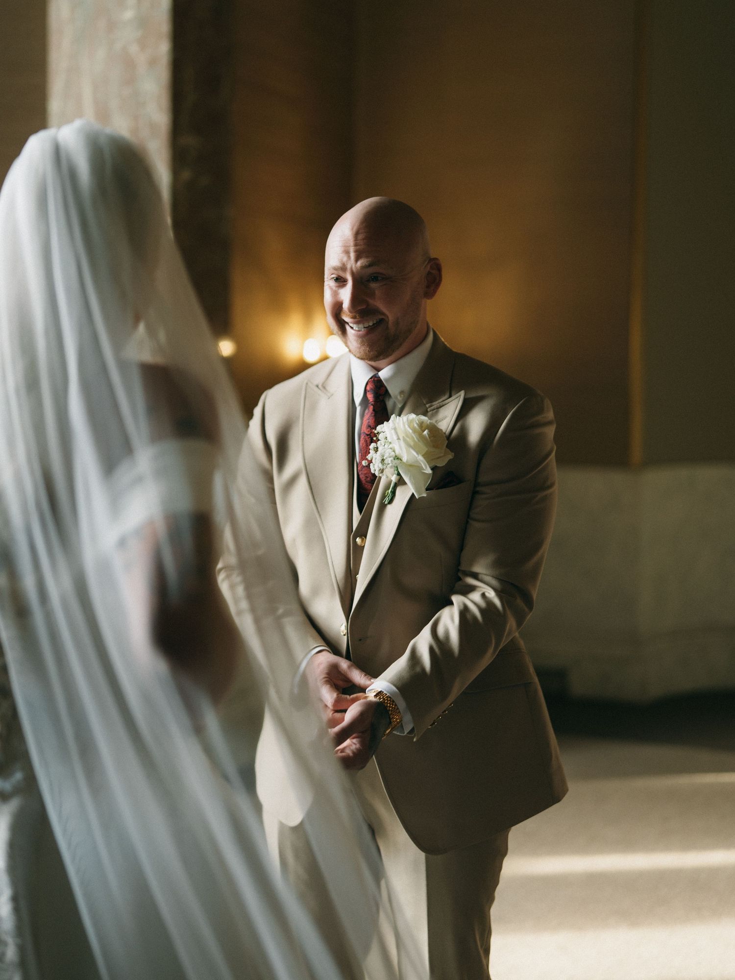 Groom in tan suit with white boutonniere smiling at bride in wedding veil during romantic ceremony.