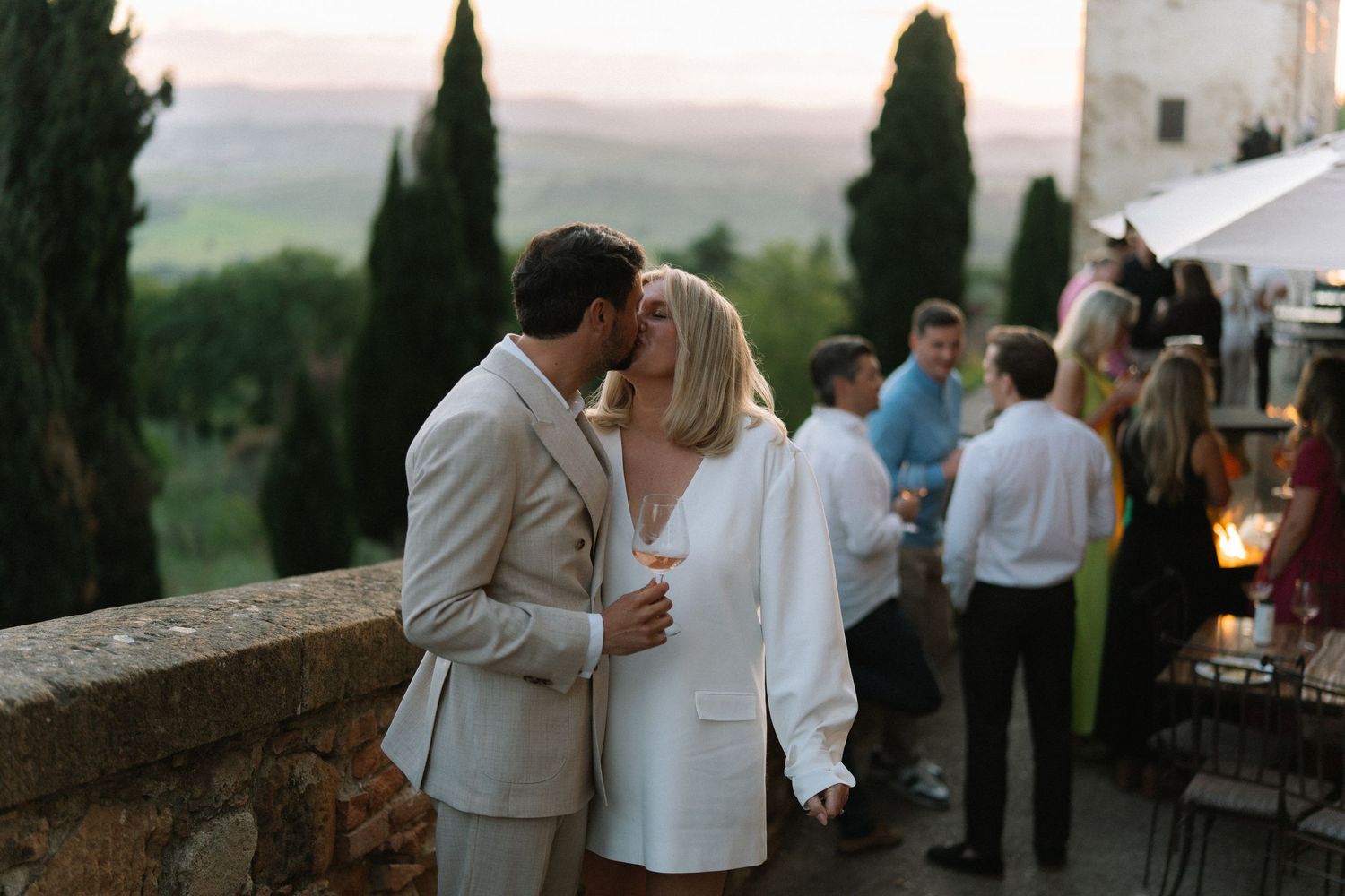 Couple shares a kiss on a terrace overlooking cypress trees and Tuscan hills at dusk.