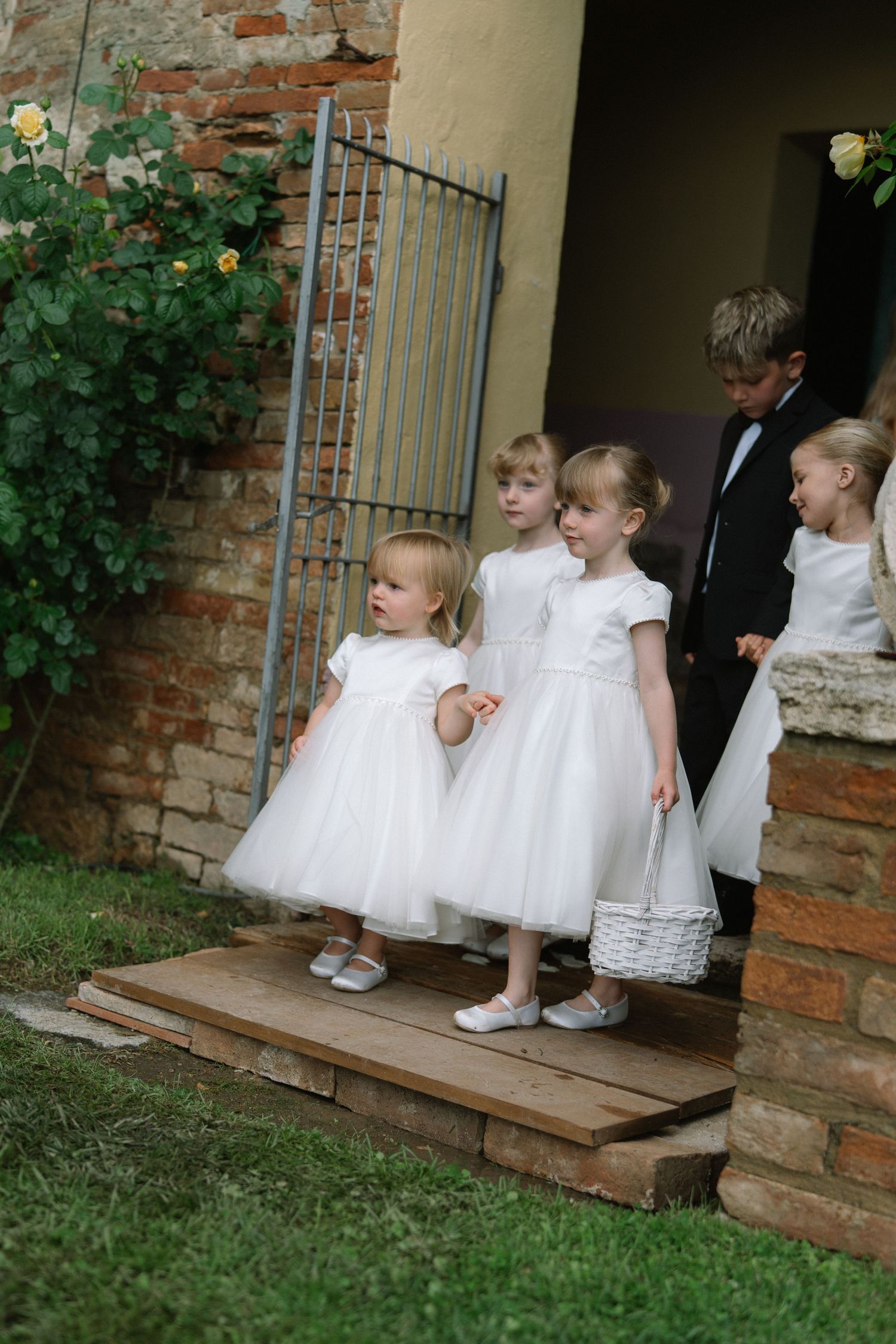 Four flower girls in white dresses and a ring bearer in a tuxedo waiting on stone steps at a rustic wedding venue.