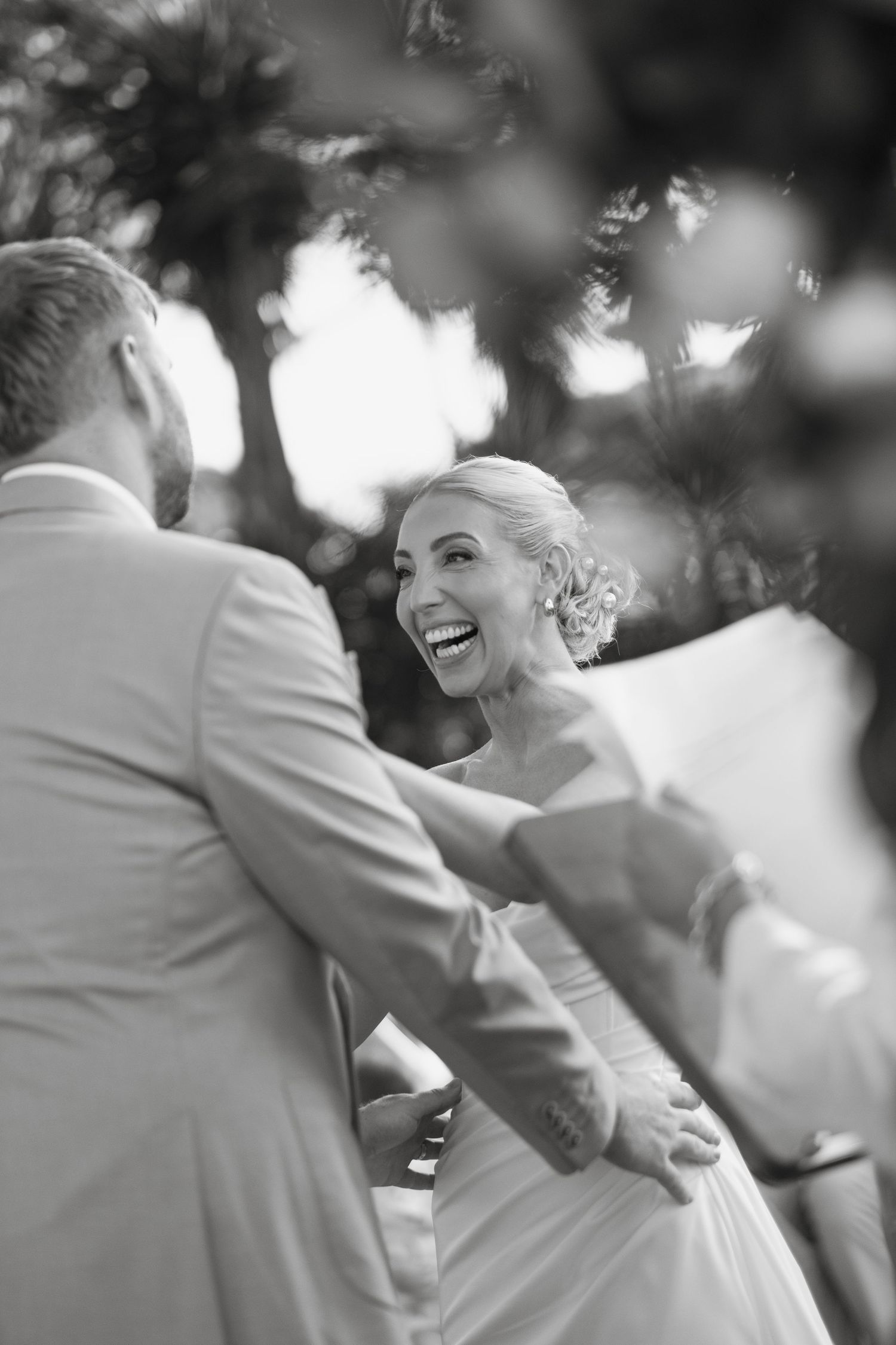 Black and white photo of a laughing bride holding hands with groom during outdoor wedding ceremony.