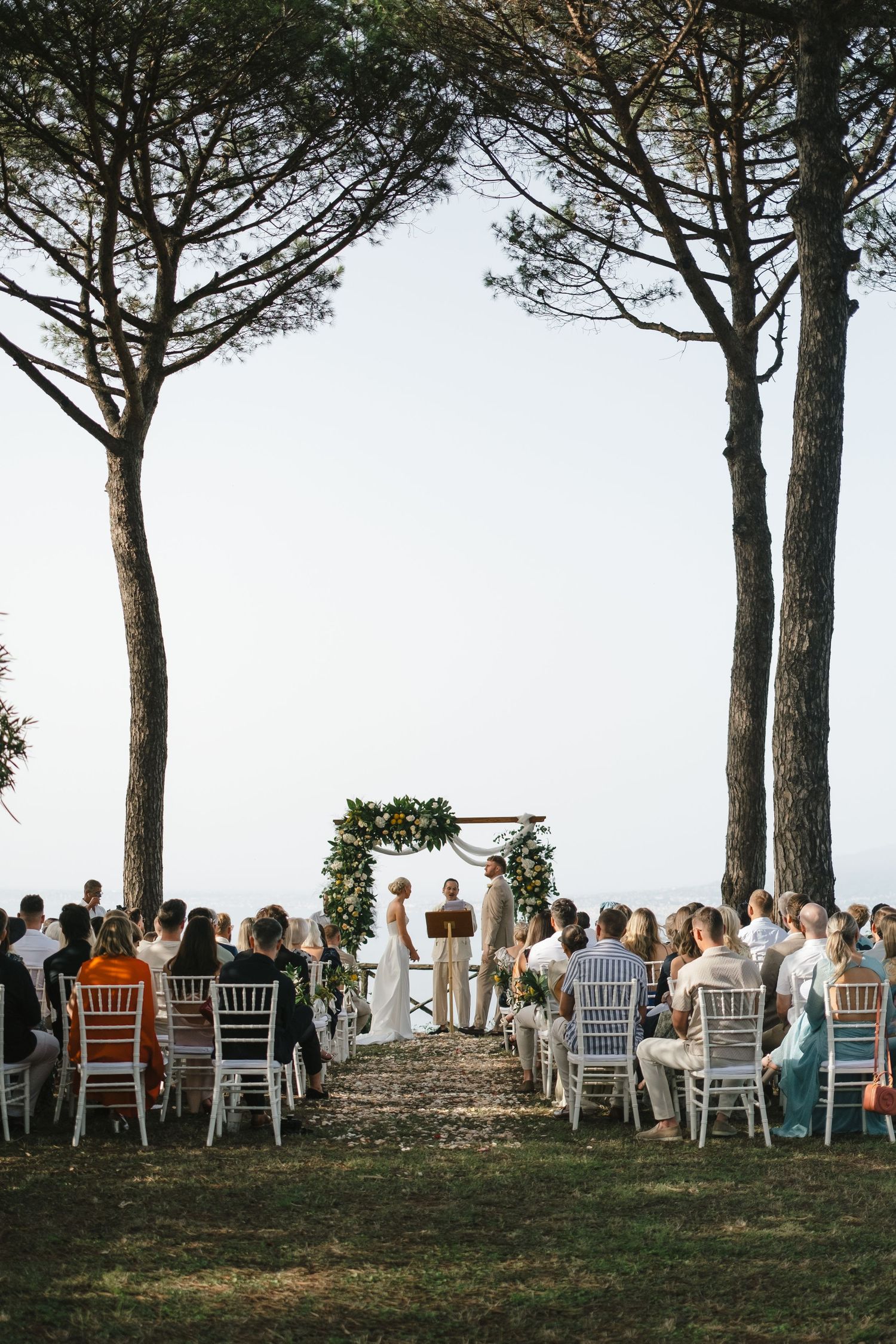 Outdoor wedding ceremony under tall pine trees with guests seated facing the couple at a floral arch.