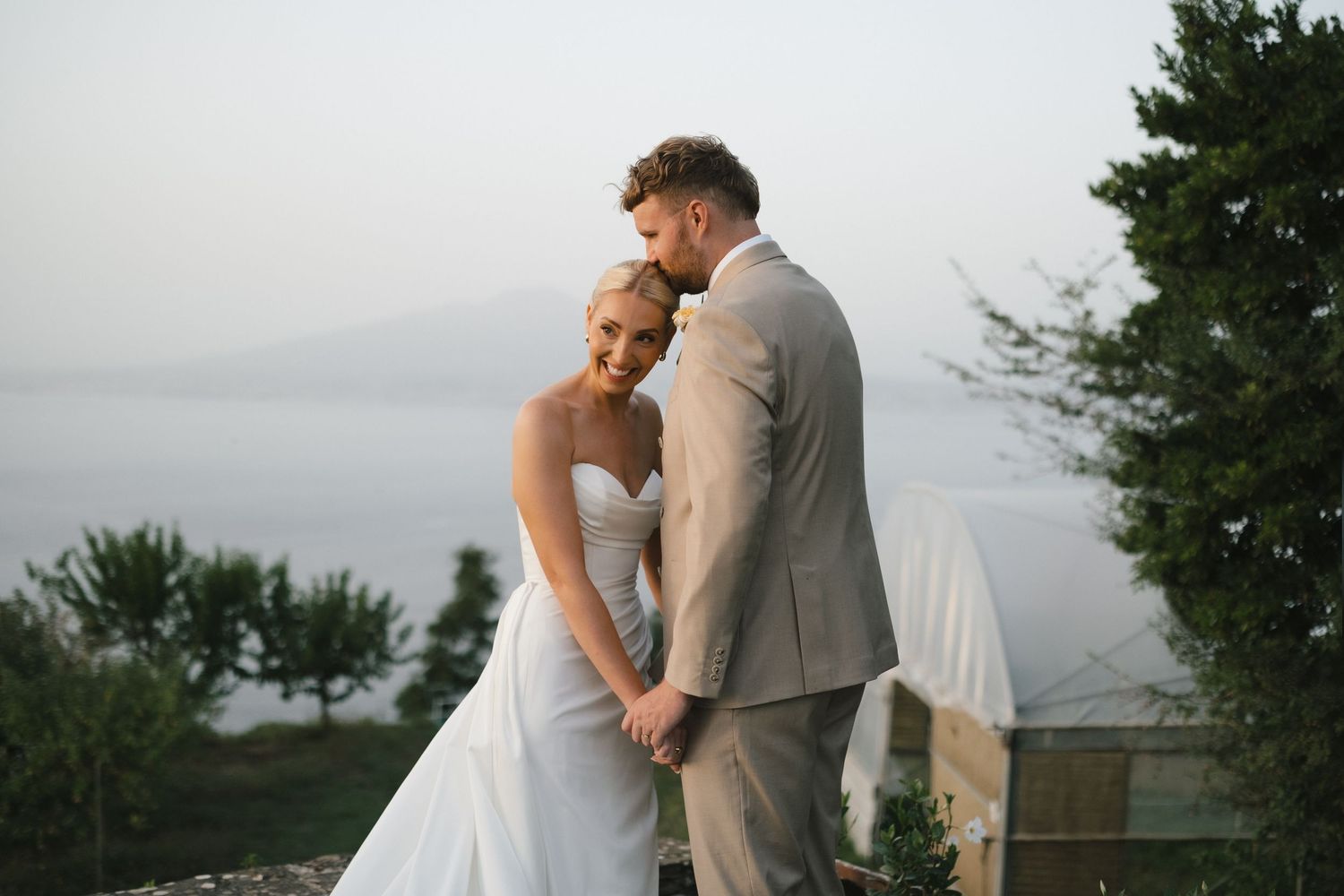 Bride and groom share tender moment with scenic coastal water views and cypress trees at sunset wedding.
