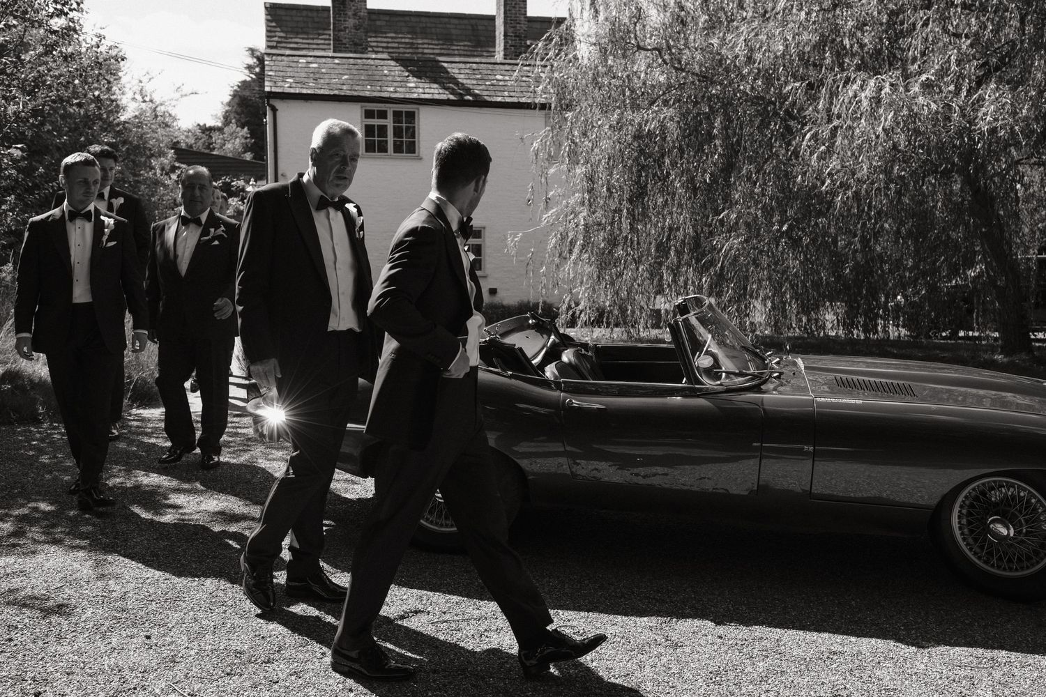 Men in tuxedos walk beside a vintage convertible car at an outdoor wedding venue with willow trees.