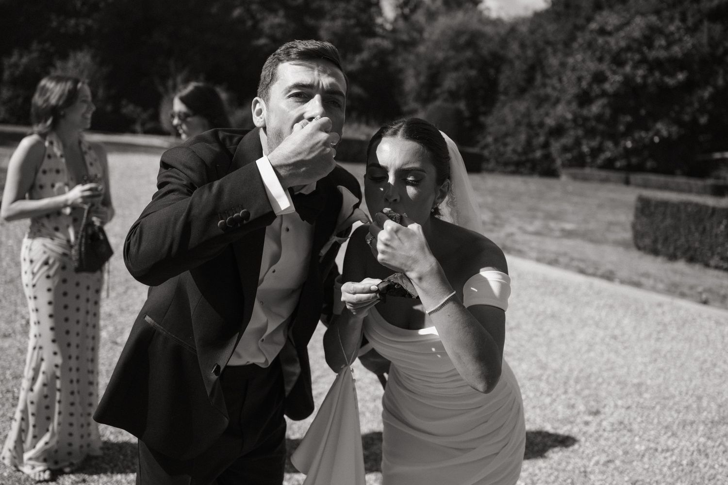 Black and white wedding photo of bride and groom playfully eating cake outdoors at elegant garden venue.
