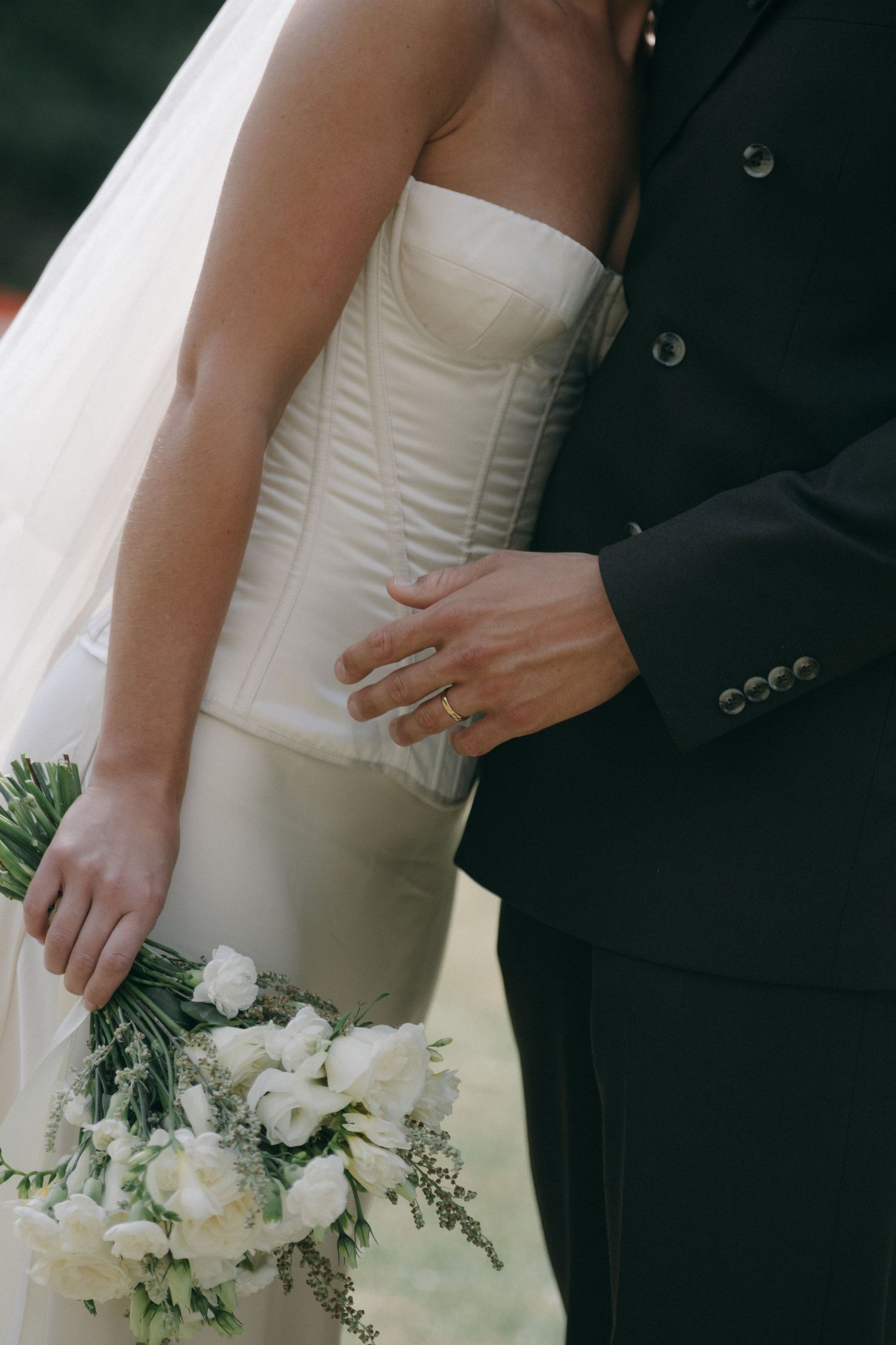 Bride in strapless white gown holding floral bouquet embraced by groom in black suit with wedding ring visible.