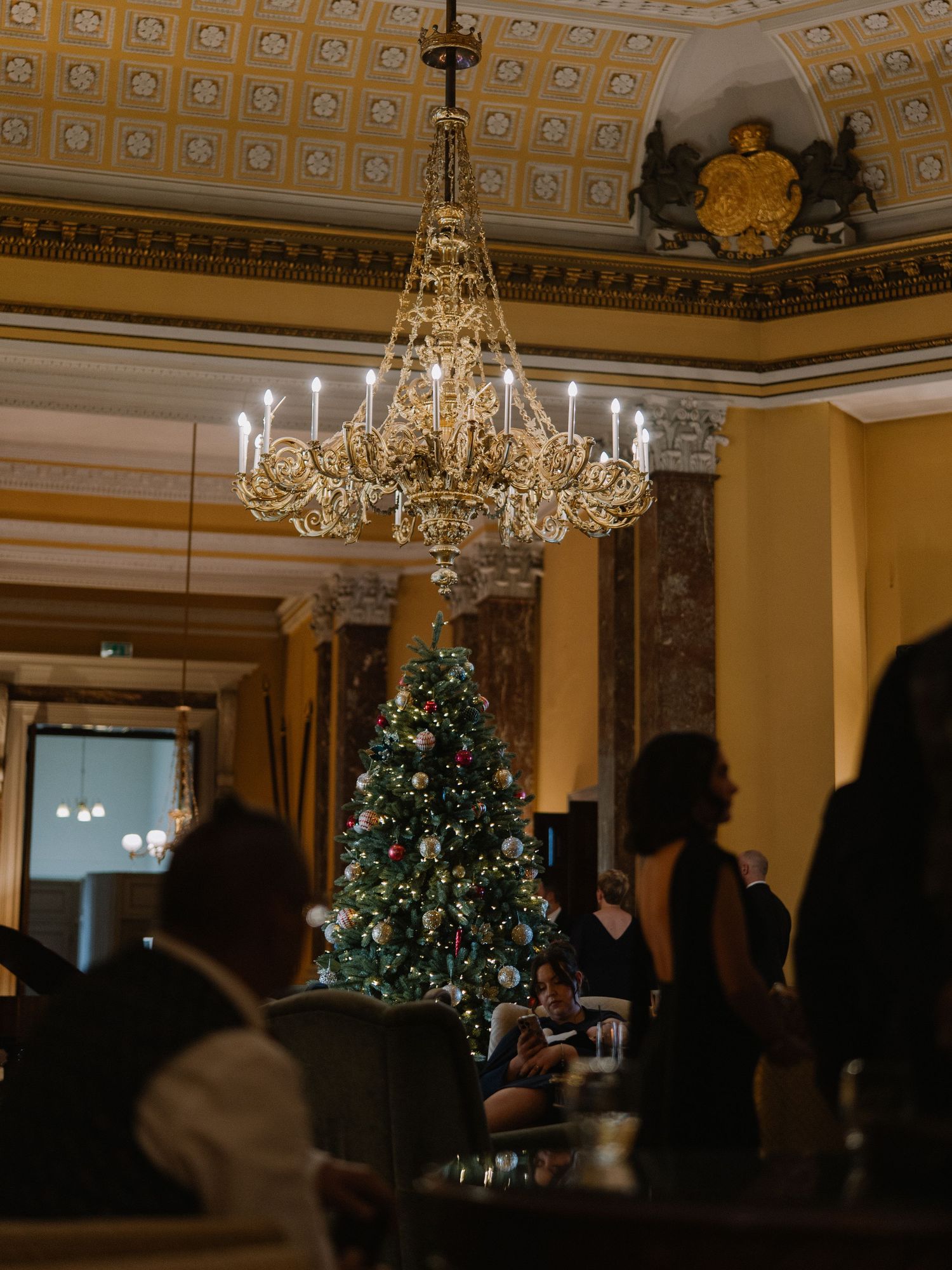 Elegant ballroom with crystal chandelier, decorated Christmas tree, and guests in formal attire during a holiday event.