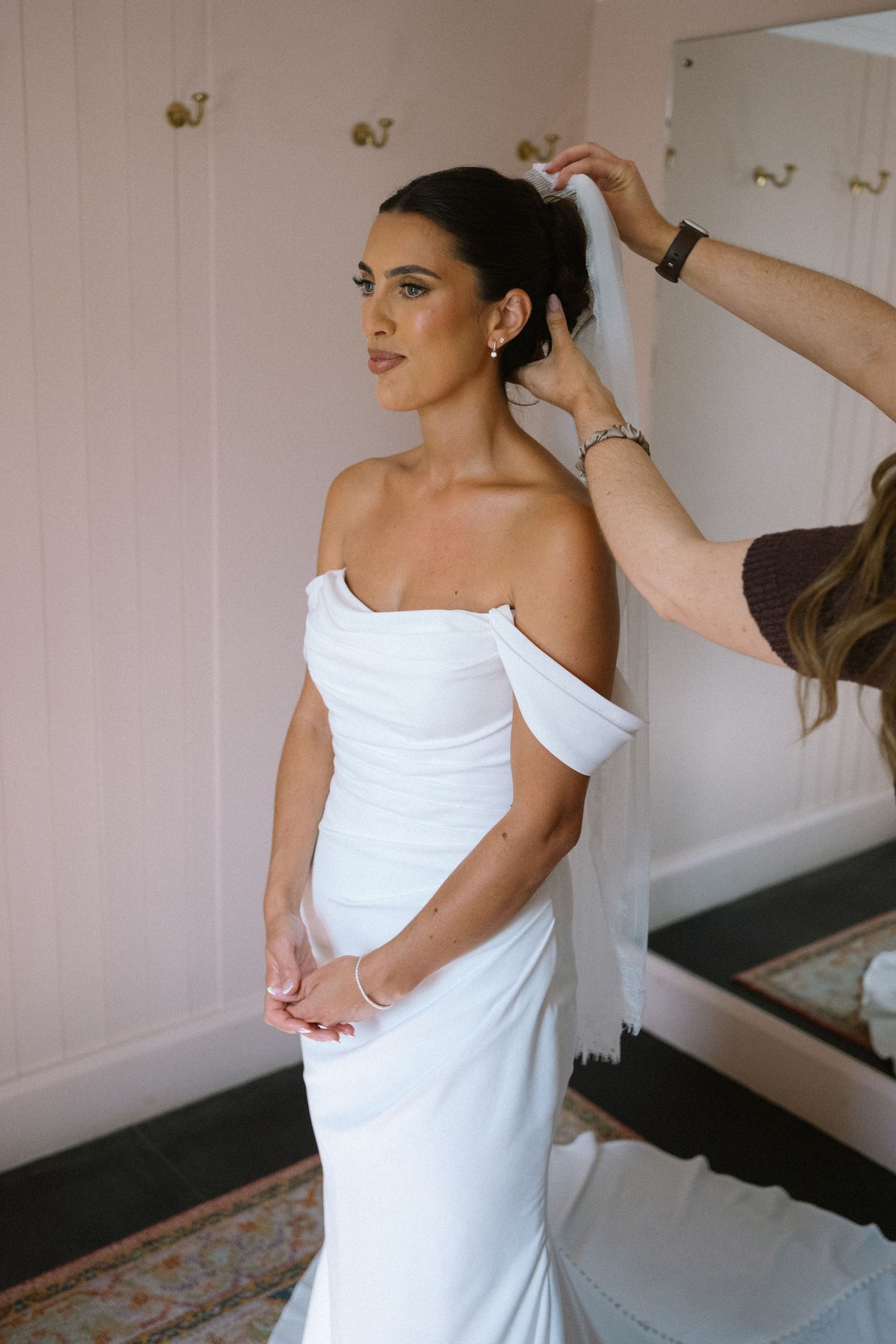 Bride in off-shoulder white gown getting veil placed by stylist in a soft pink room.