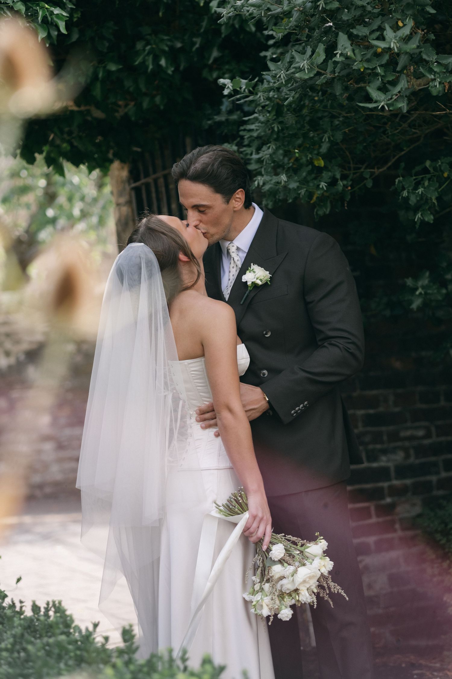 Bride and groom share a romantic kiss outdoors, bride holding white bouquet wearing veil, groom in dark suit with boutonniere.