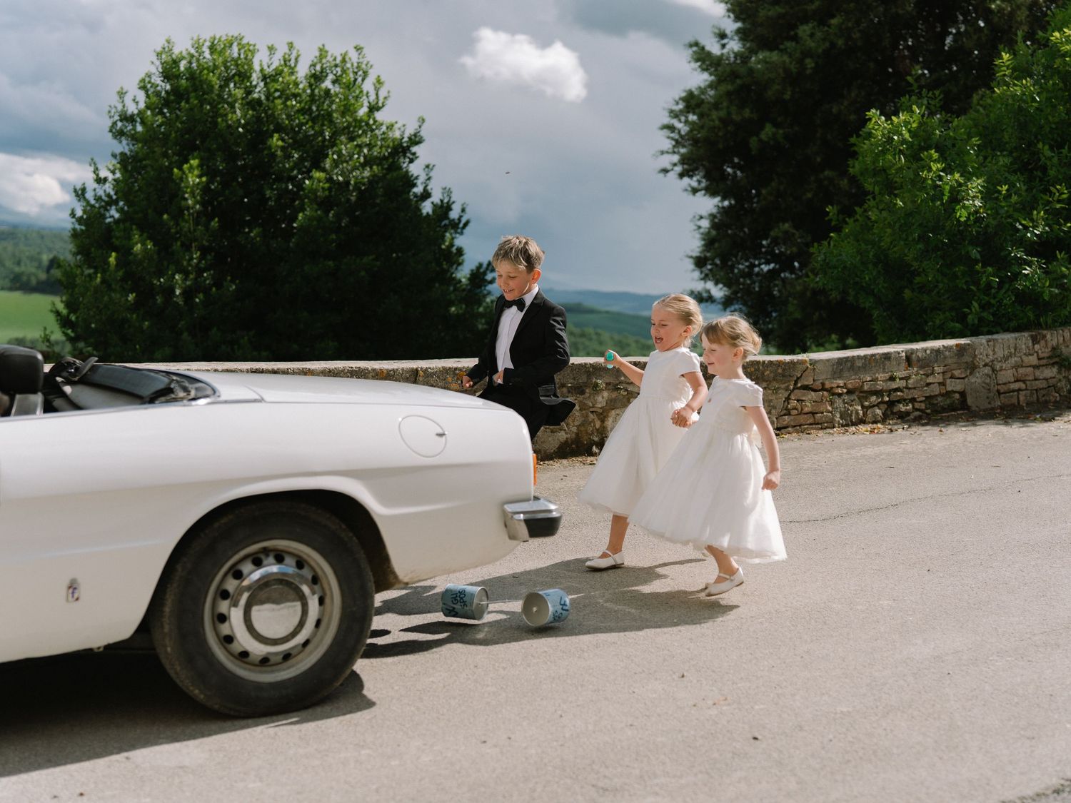 Children in formal wear pose beside a vintage white convertible car at an outdoor wedding celebration.