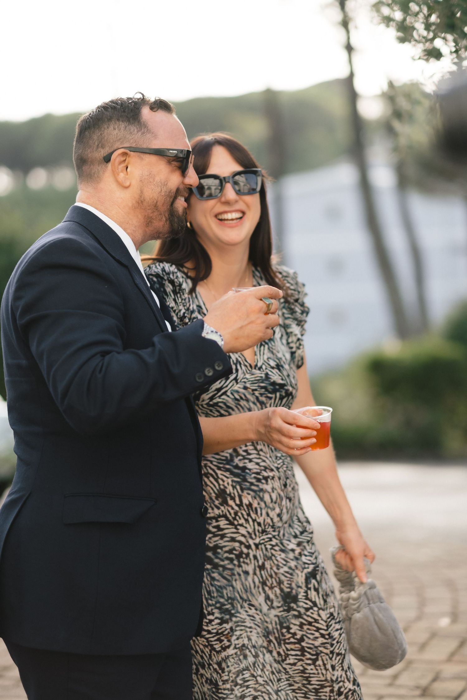 Couple in sunglasses smiling and holding drinks at an elegant outdoor wedding reception.