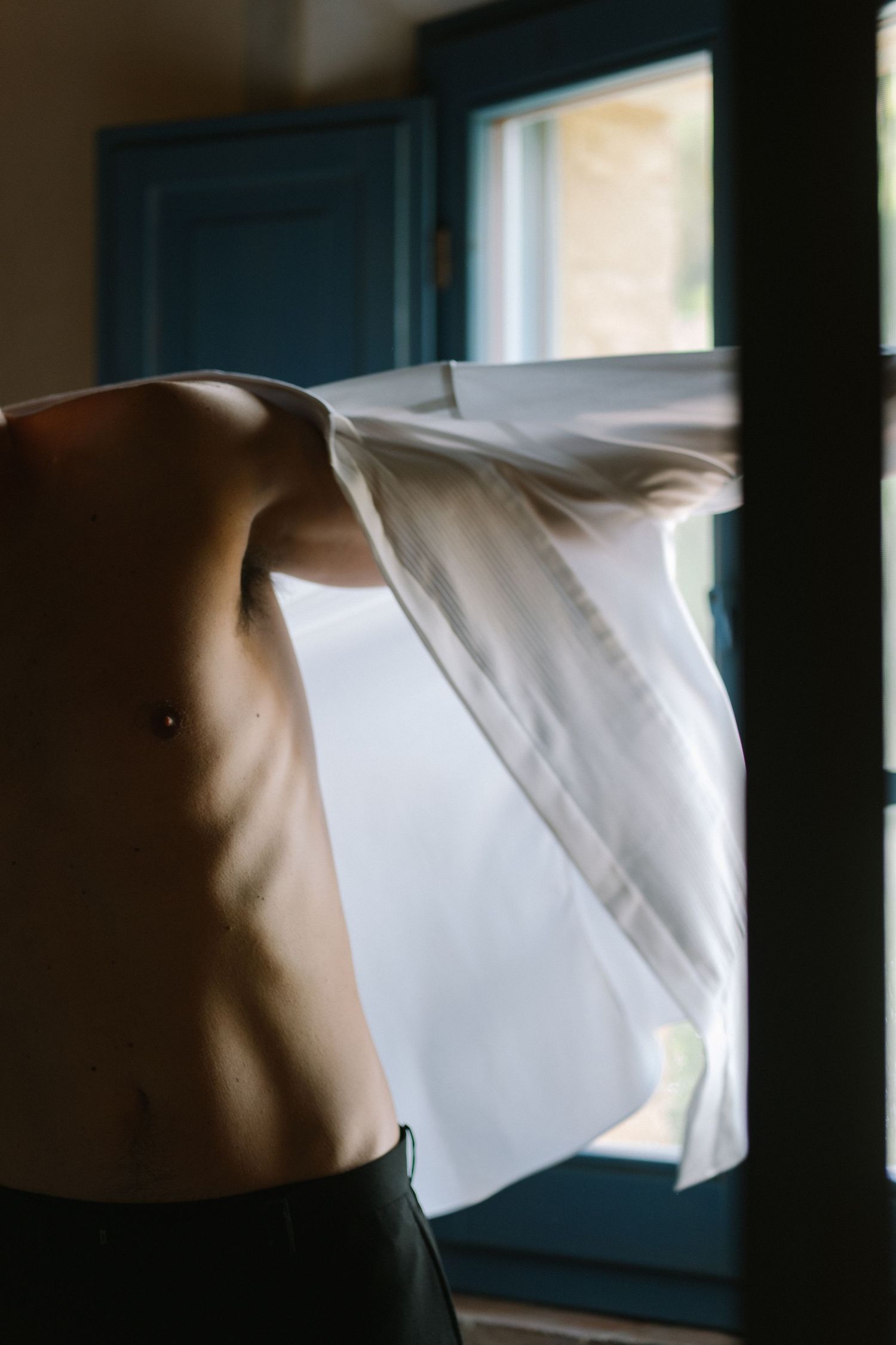 Groom putting on a white dress shirt backlit by light from a blue-shuttered window.