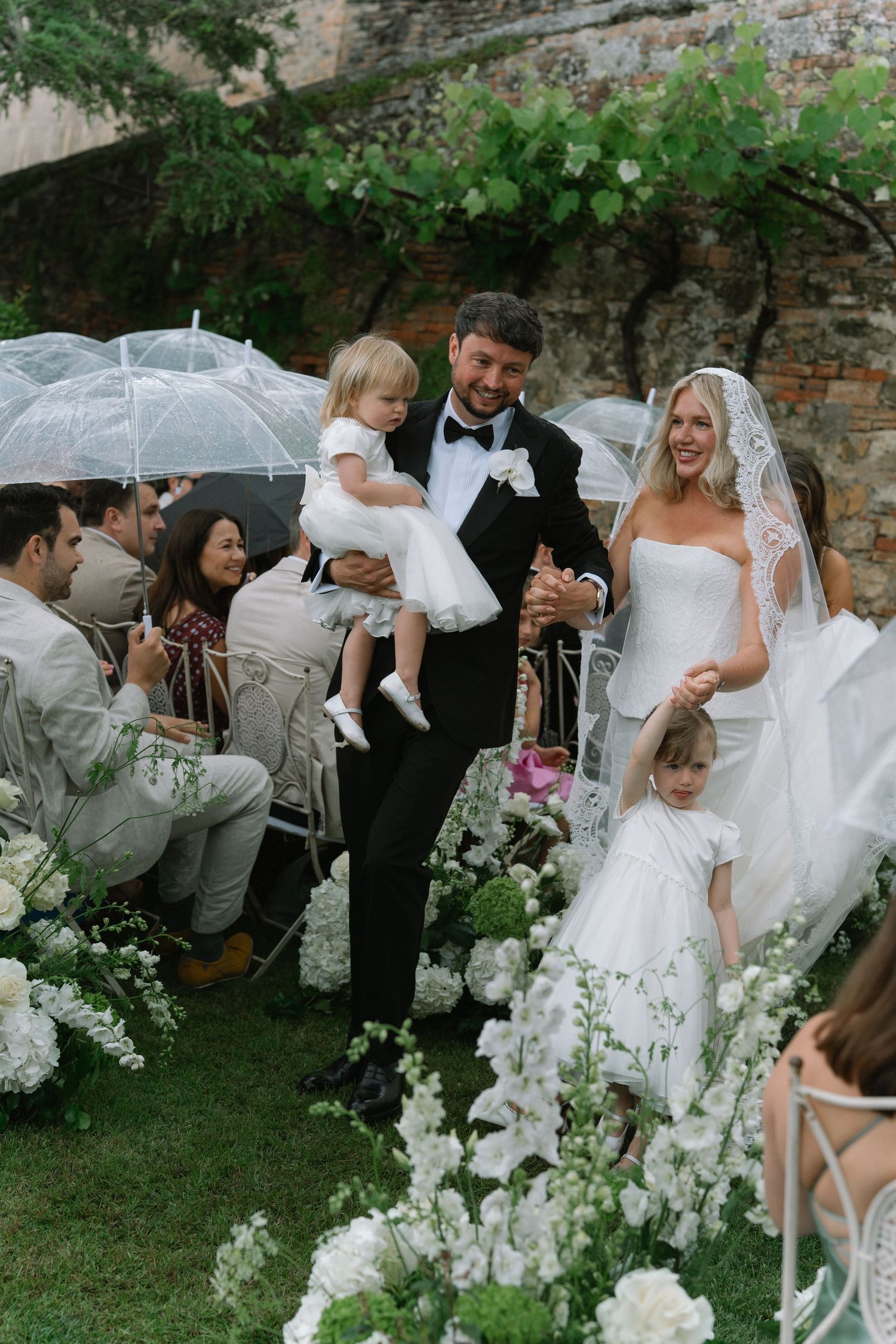 Groom carries flower girl while walking down the aisle with bride after outdoor wedding ceremony.