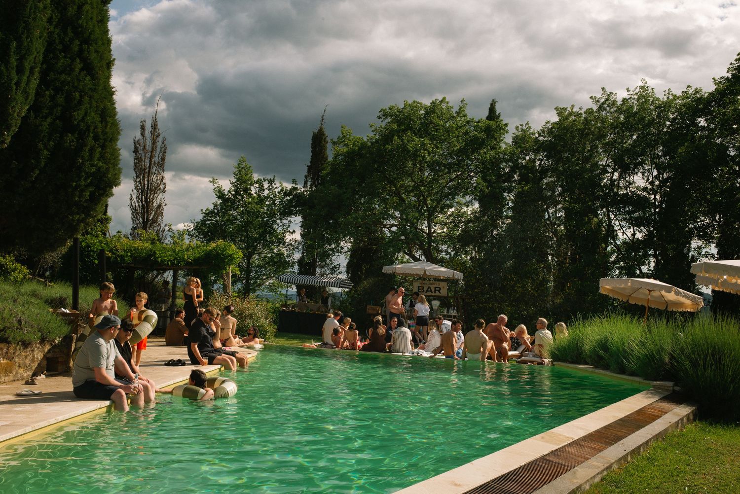 Guests relaxing by a scenic Tuscan villa pool surrounded by lush greenery under dramatic cloudy skies.