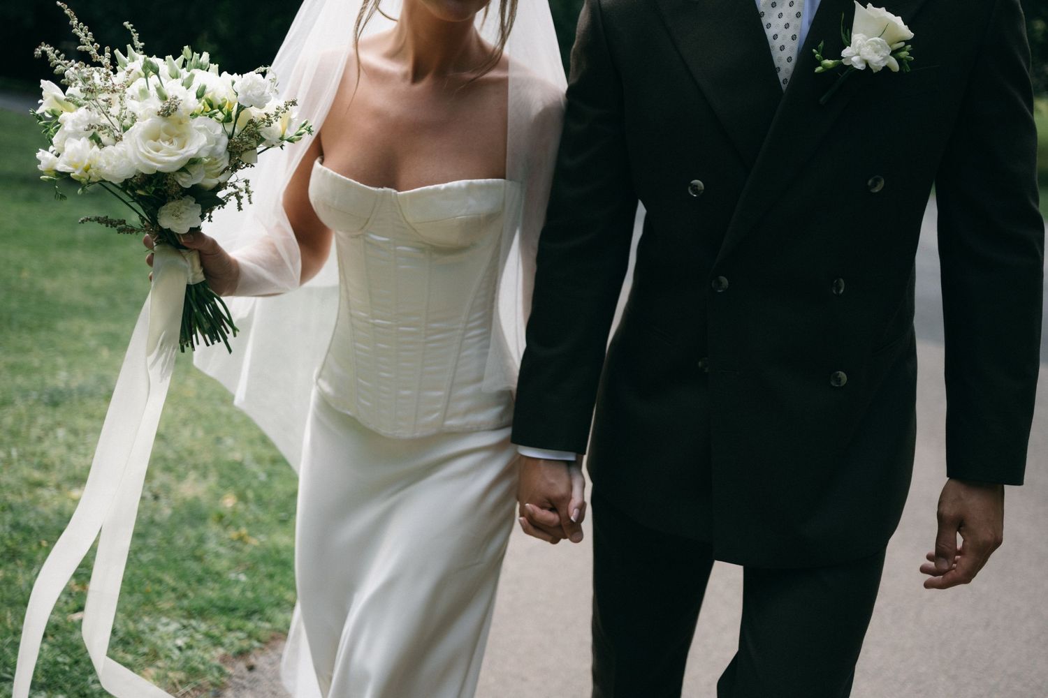 Bride in white corset gown holding bouquet walks hand in hand with groom in black suit on garden path.