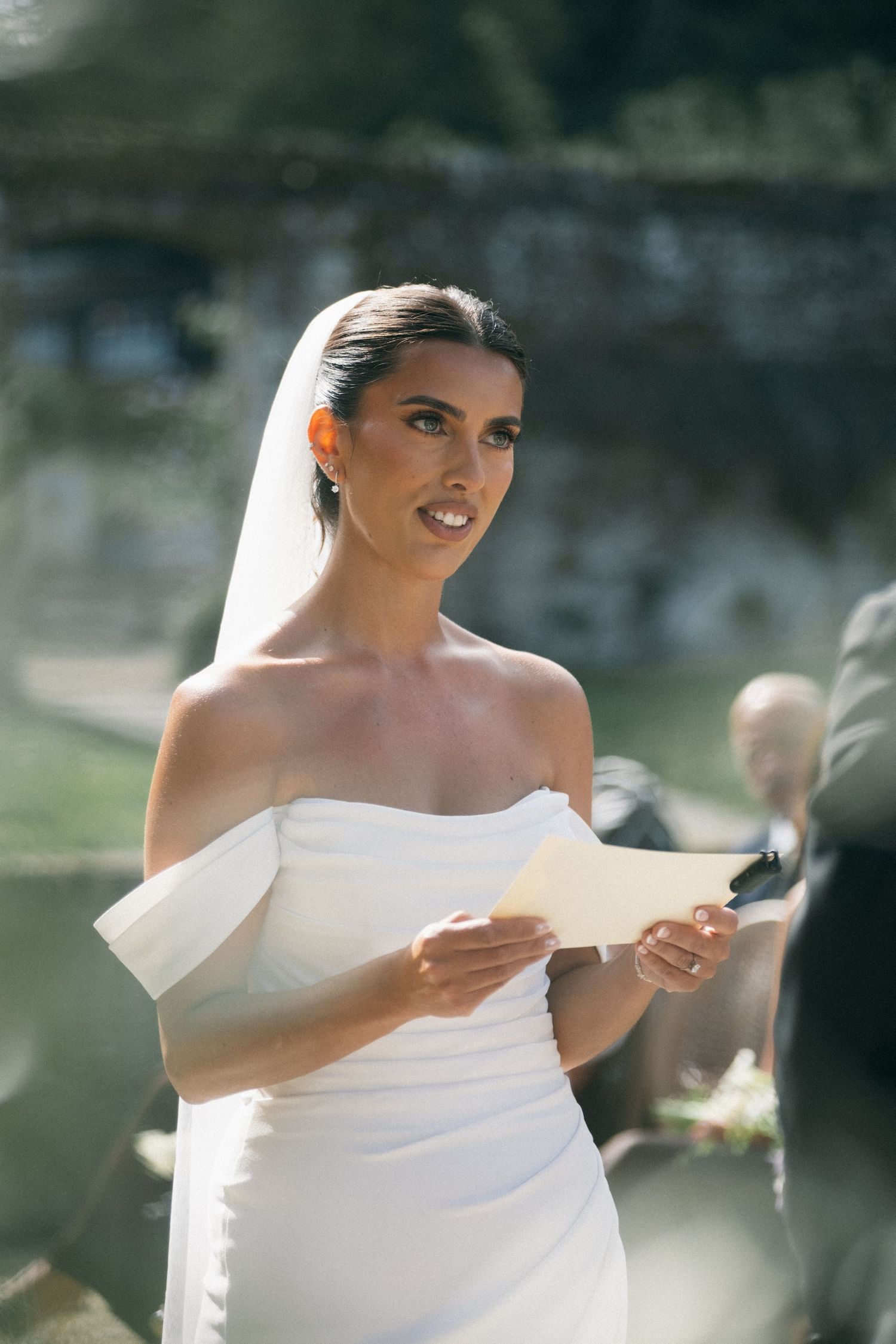 Elegant bride in off-shoulder white gown and veil reads wedding vows from a card during outdoor ceremony.