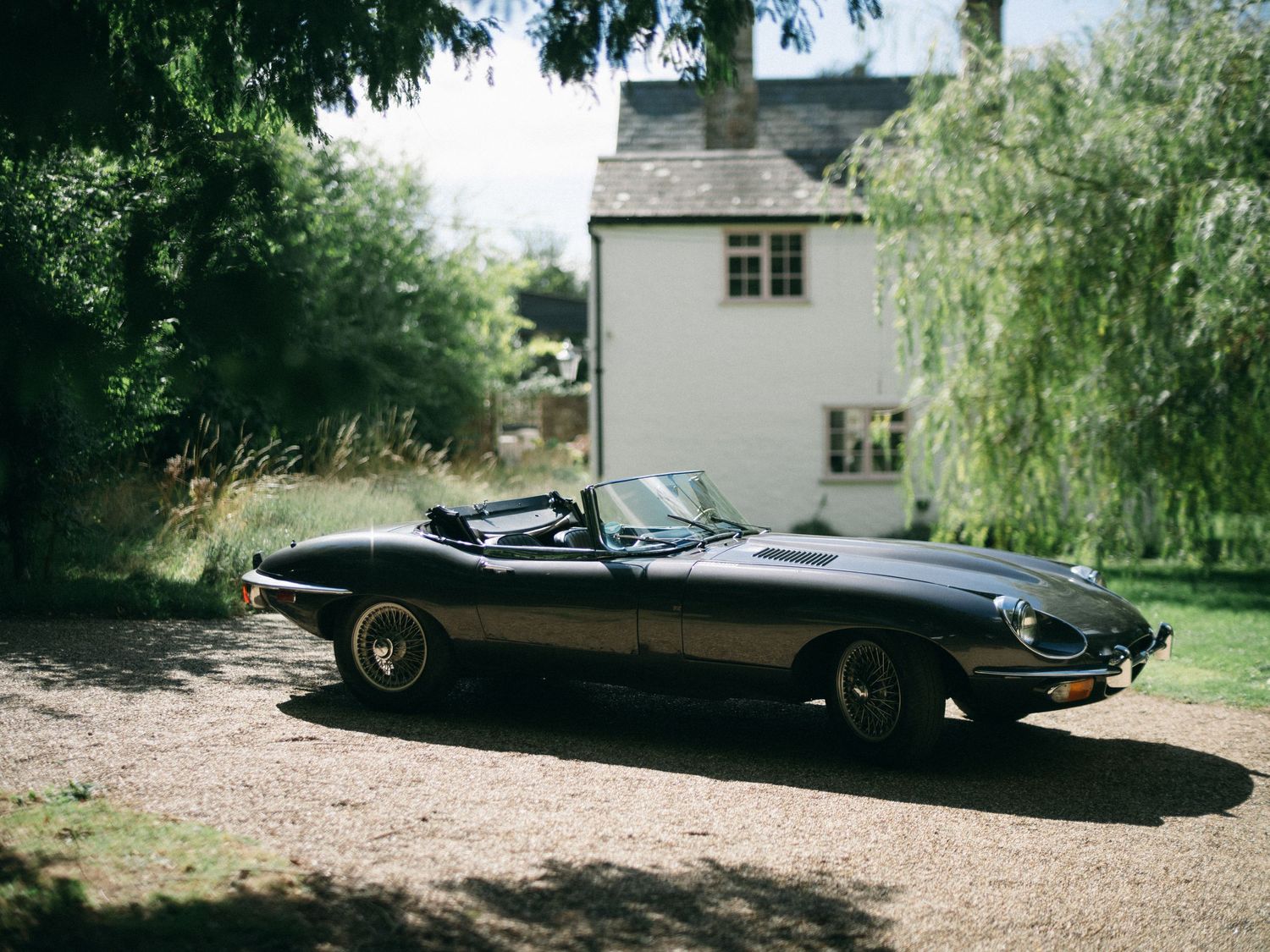 Black vintage Jaguar E-Type convertible parked on gravel driveway beside a white country cottage surrounded by lush trees.