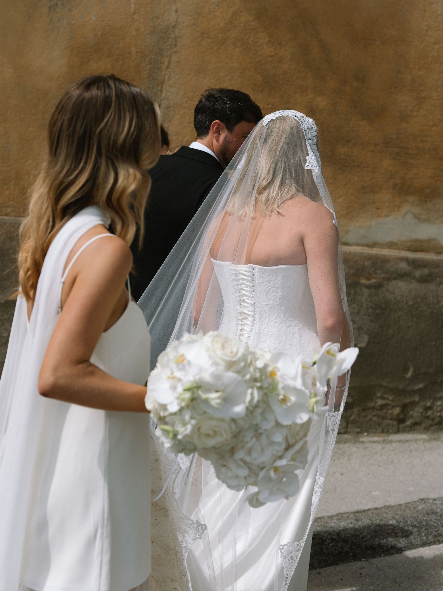 Bride in a strapless gown holding a white bouquet walking with bridesmaids along a rustic Italian stone wall.