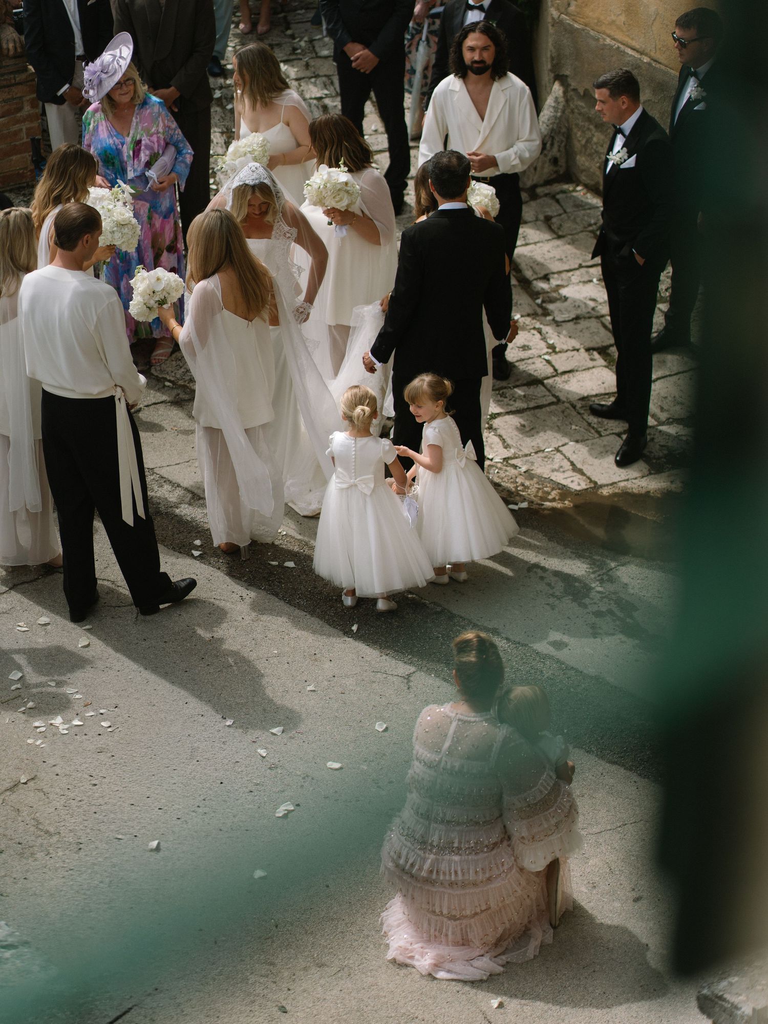 Wedding party gathers on cobblestone street with flower girls, bridesmaids in white, and guests mingling.