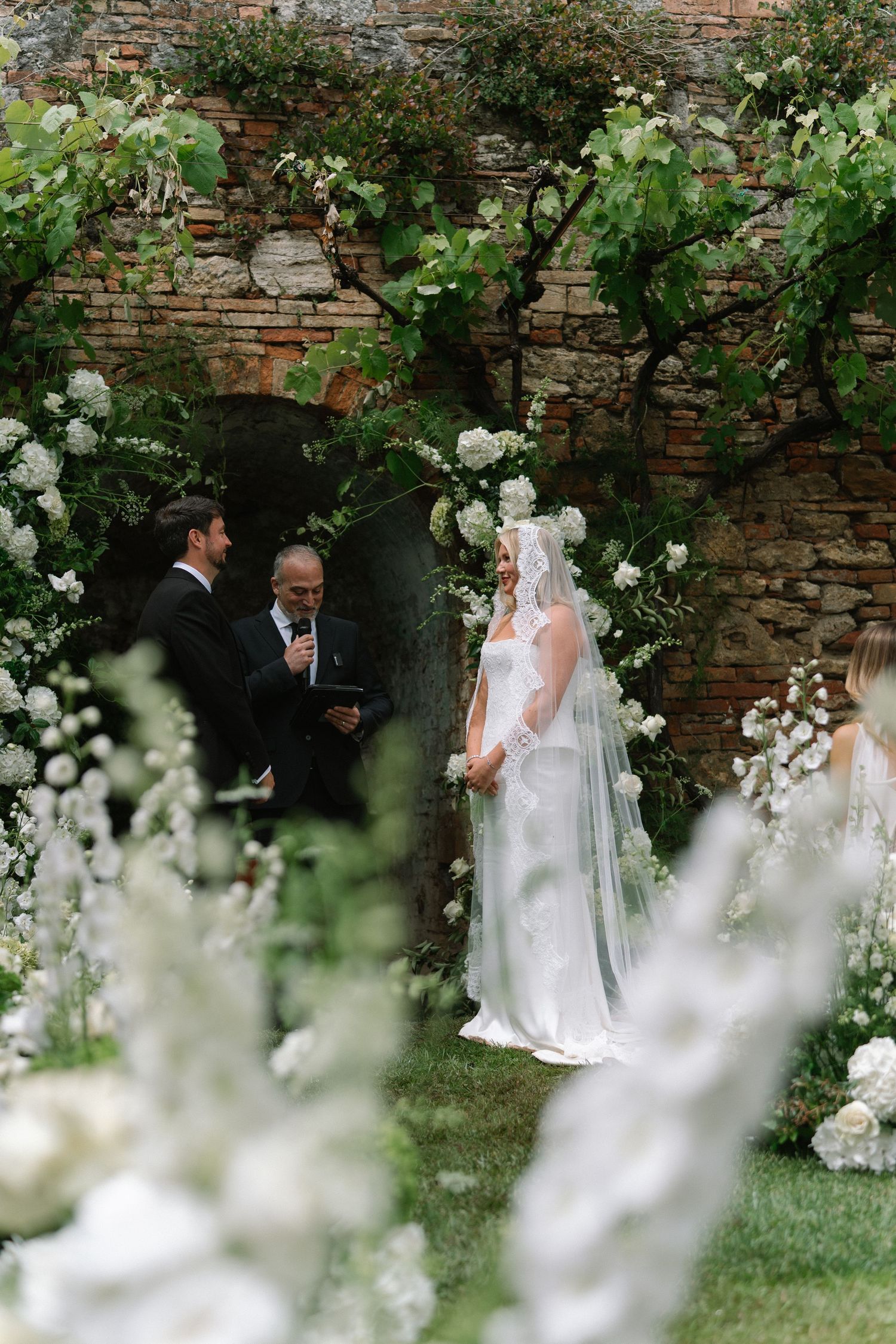 Bride smiles at groom during outdoor wedding ceremony framed by white hydrangeas and greenery.