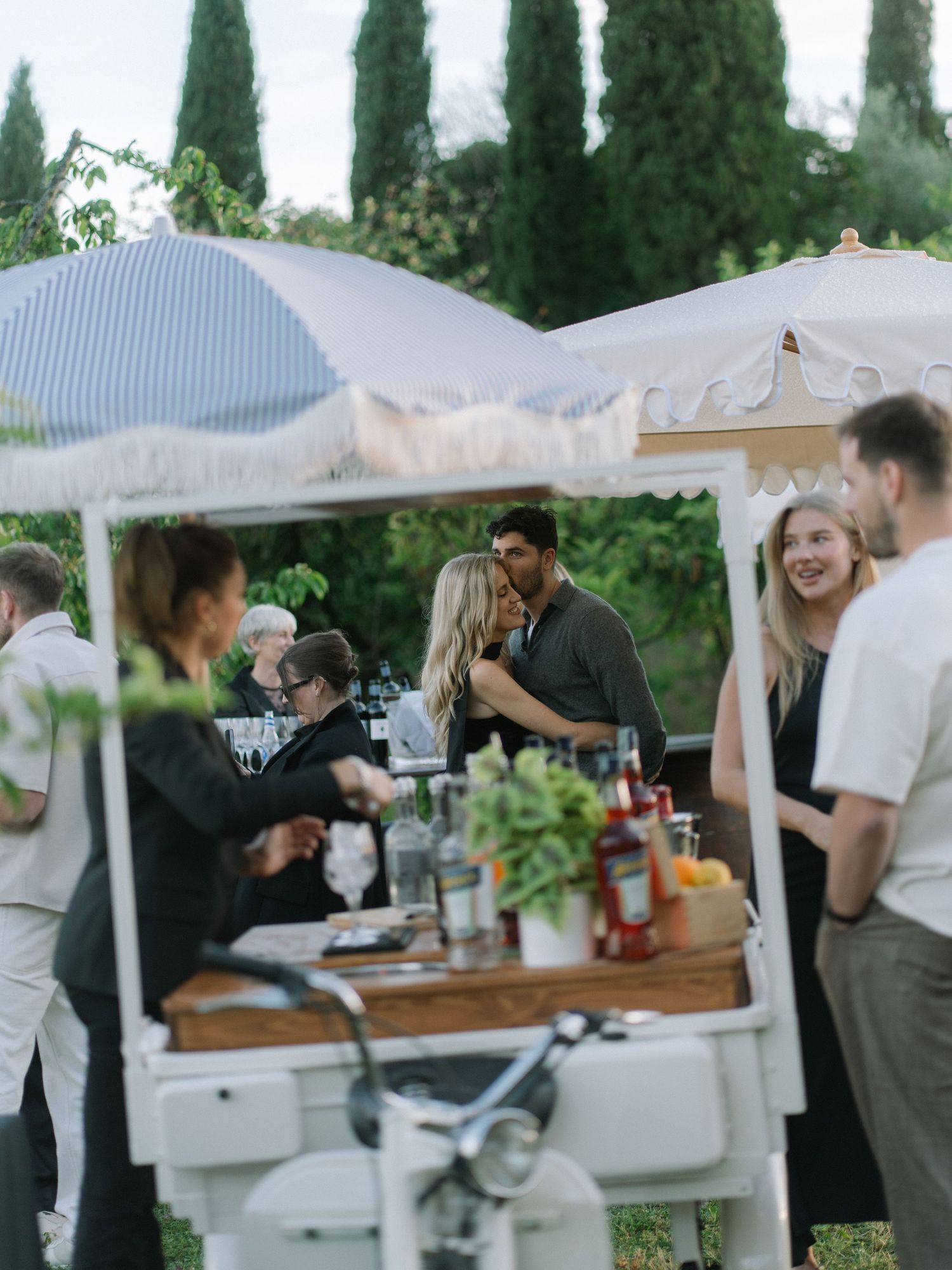 Wedding guests mingling around a vintage white bar cart with cypress trees in the background at dusk.